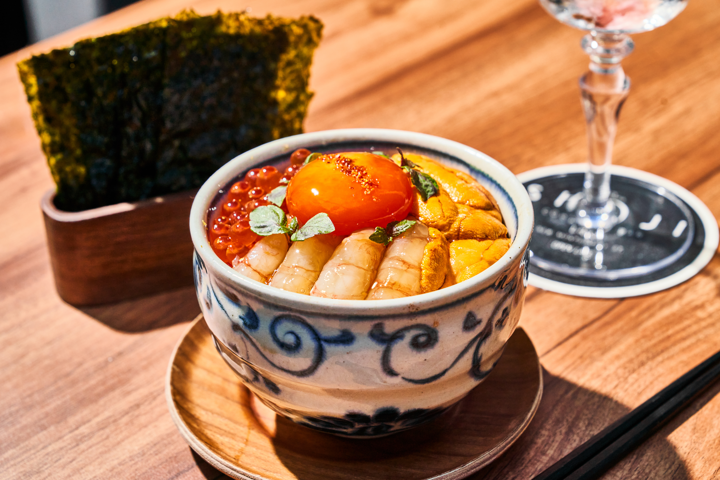 A decorative bowl contains seafood, including shrimp, sea urchin, and salmon roe, topped with herbs and an egg yolk. Seaweed sheets and a drink are beside it.