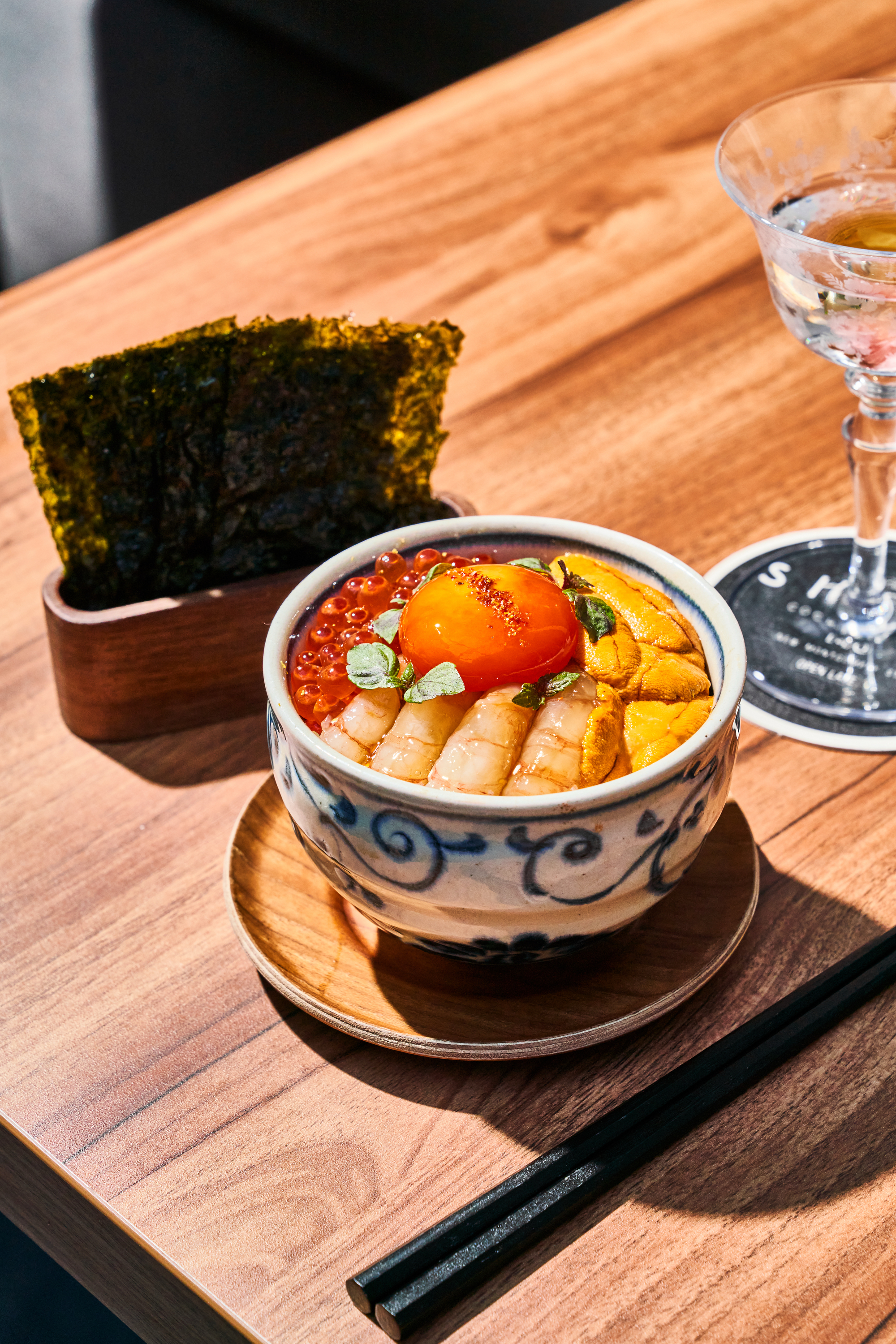 A bowl of sushi rice is topped with shrimp, sea urchin, salmon roe, and a raw egg yolk, beside seaweed sheets and a cocktail on a wooden table.