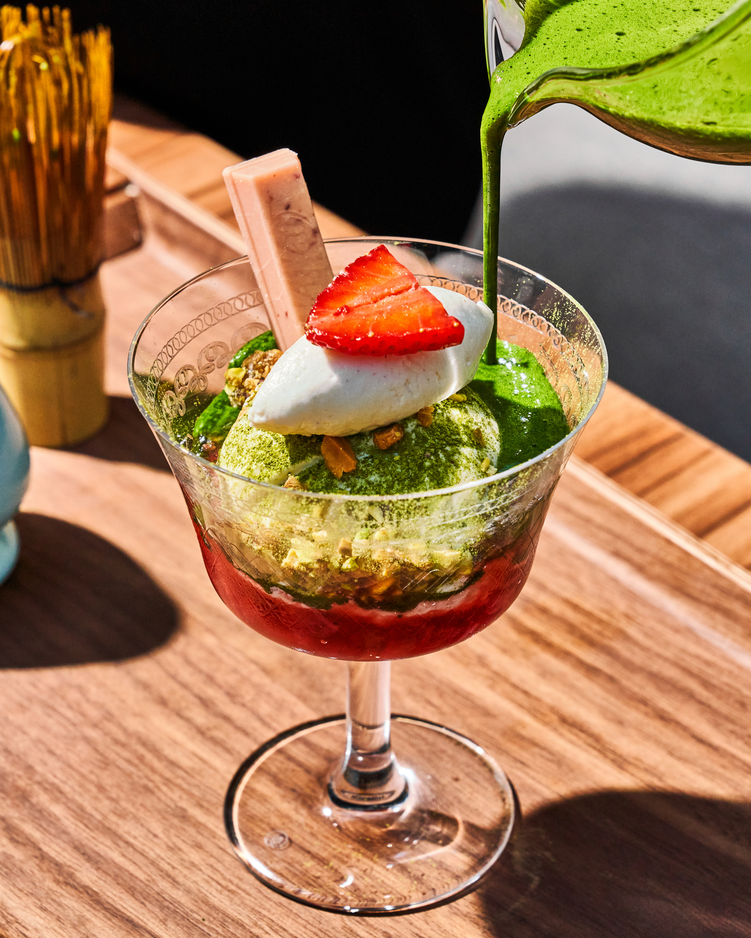 A dessert glass with green matcha being poured, containing ice cream, nuts, strawberry, whipped cream, and a rectangular pink wafer on top.