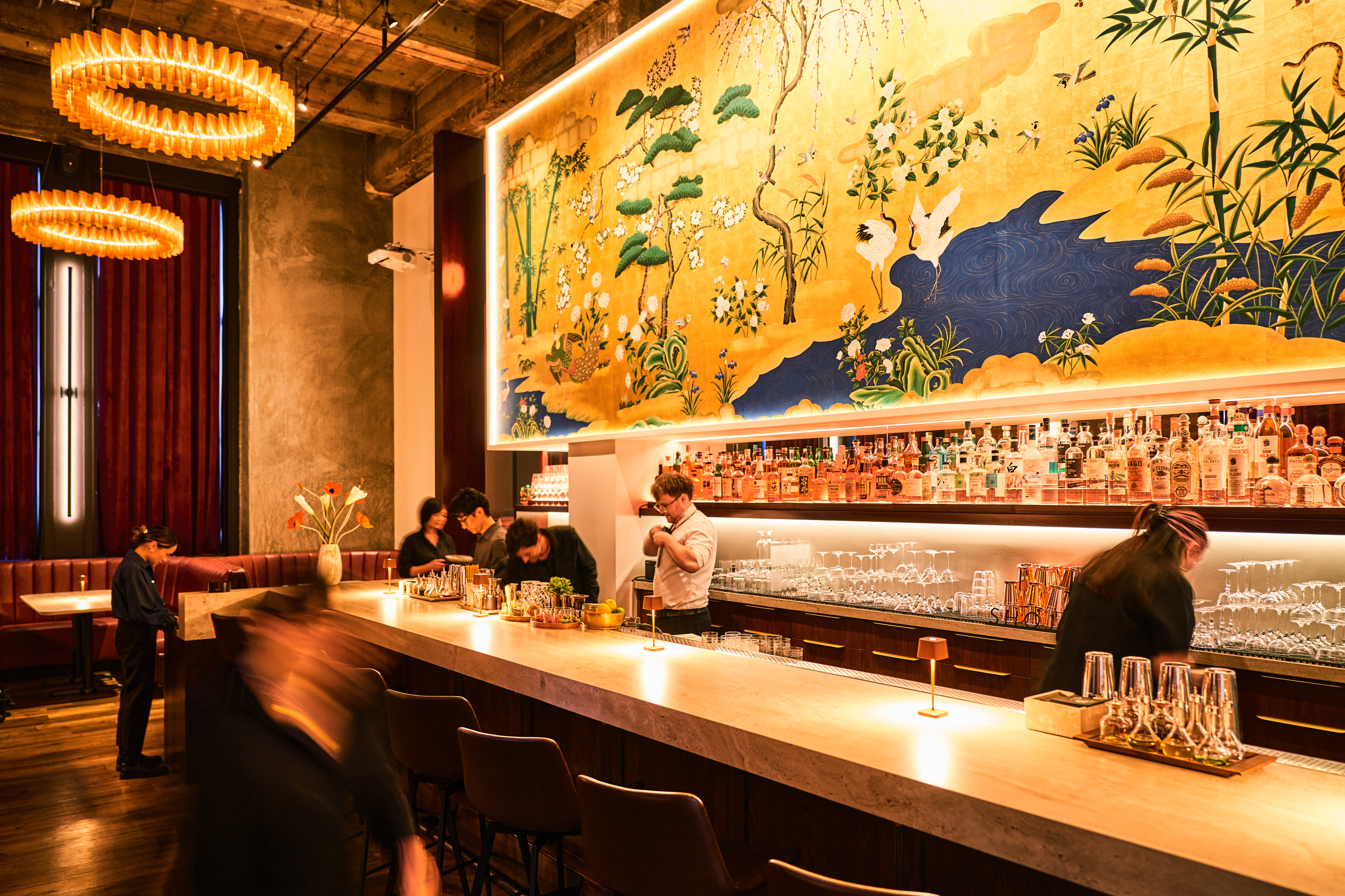 A vibrant bar features a large, colorful mural above, depicting nature scenes with birds and trees. Several people are working behind a sleek, well-stocked bar.