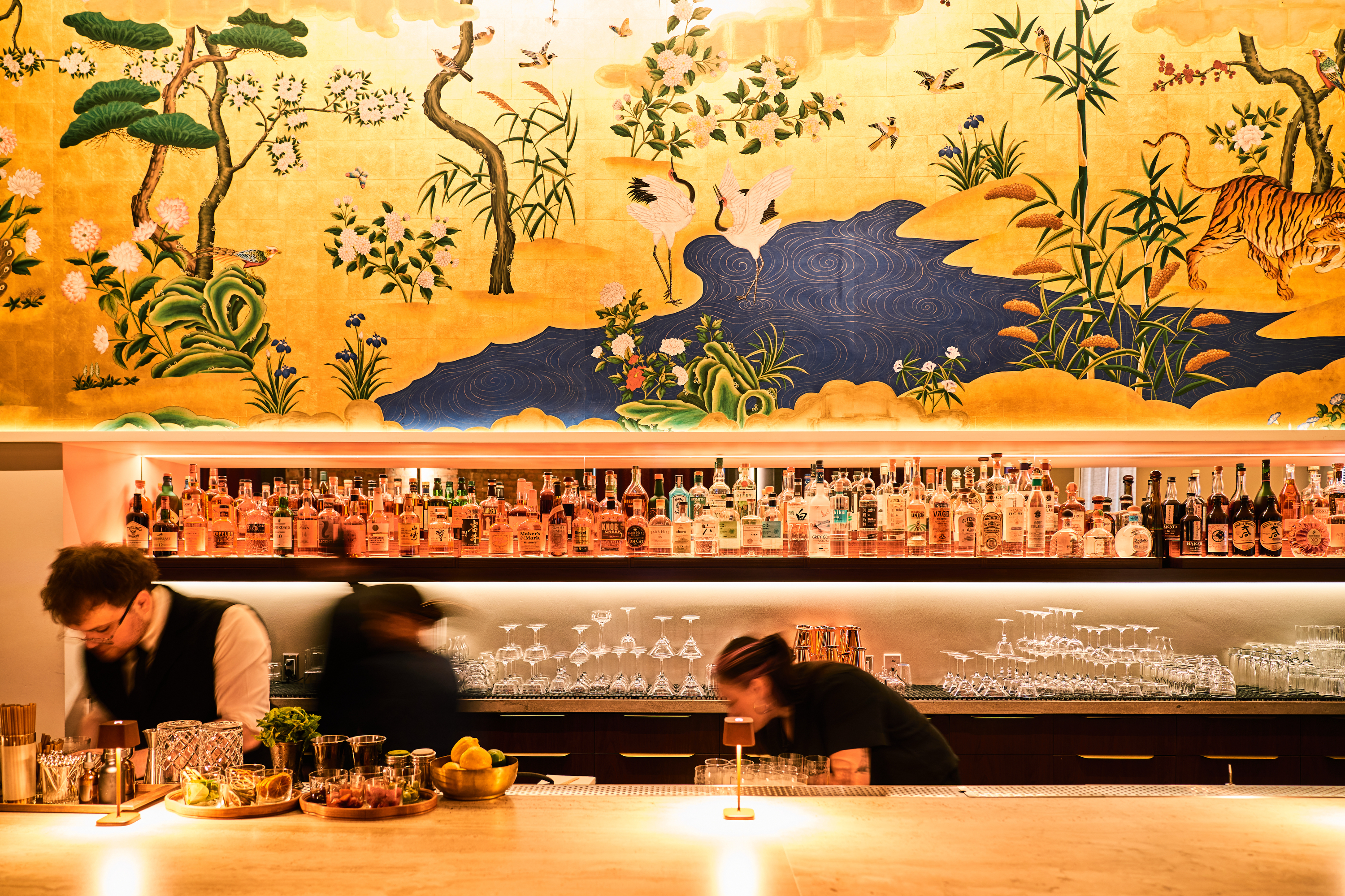 A vibrant bar scene features three bartenders preparing drinks. Behind them is an ornate mural with a nature motif. Shelves hold numerous bottles and glasses.