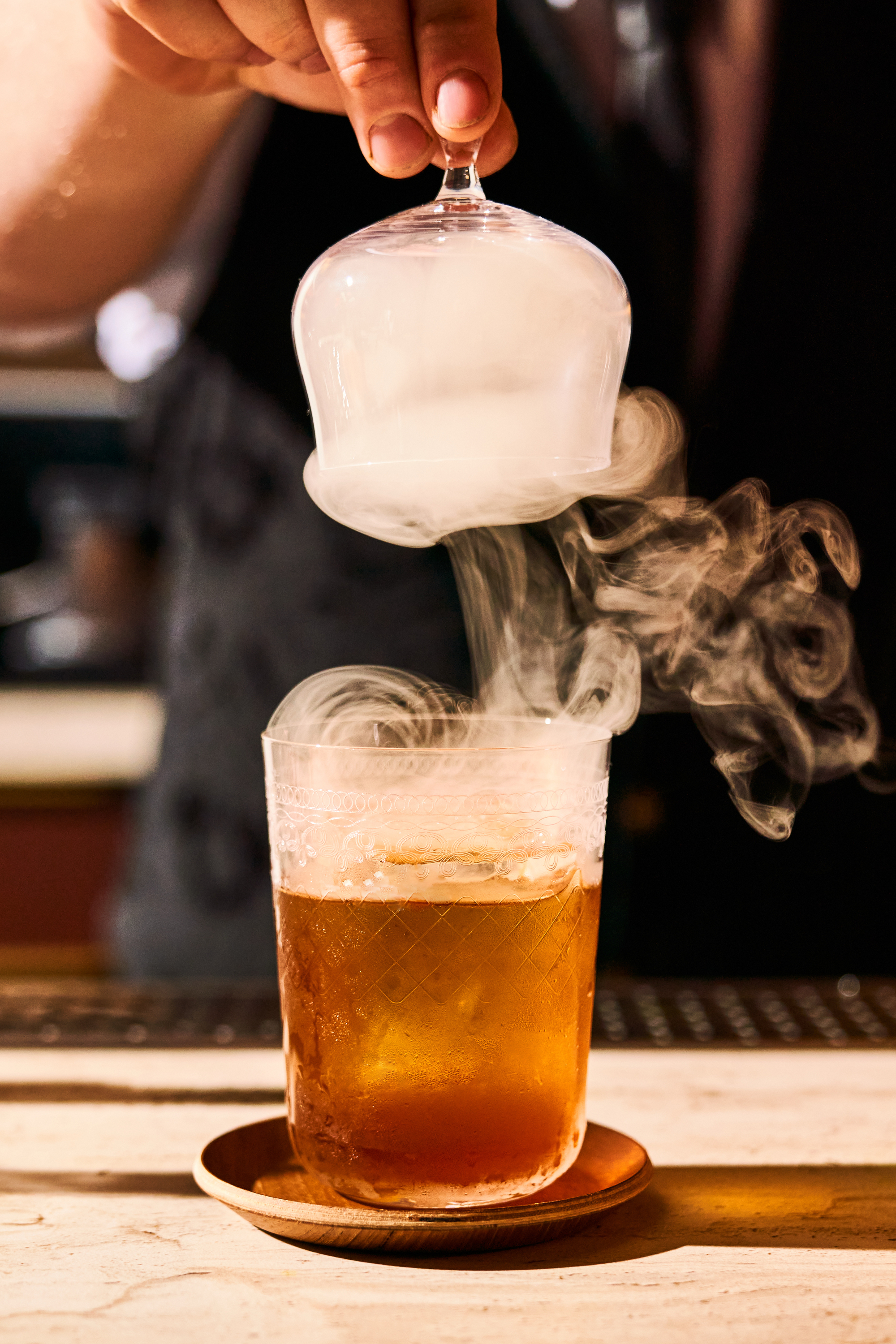 A hand lifts a glass cloche releasing smoke from an amber-colored drink in a glass with ice on a wooden coaster.