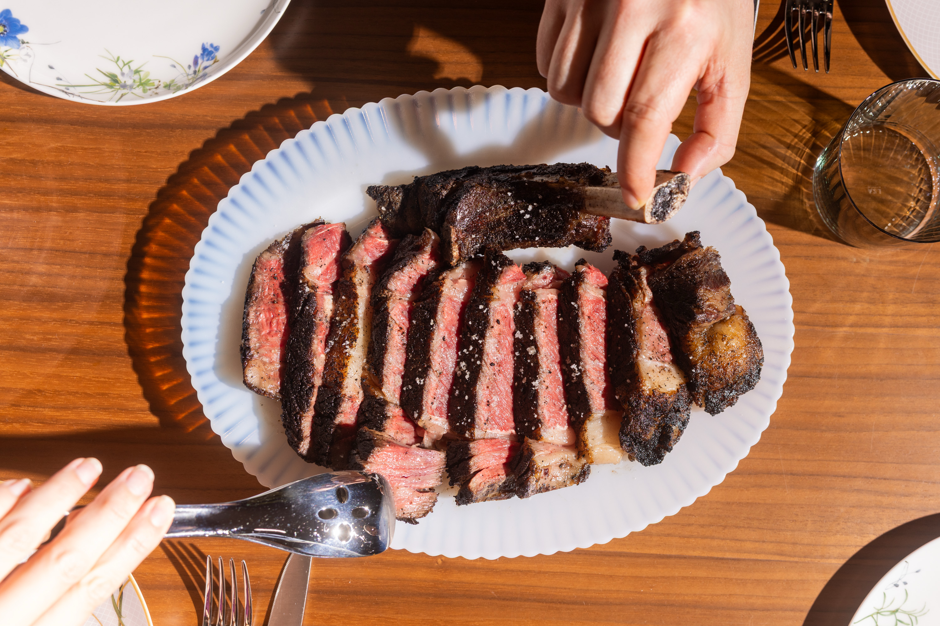 A sliced, seasoned steak is displayed on a white plate, with a hand reaching for a bone. Forks and a glass are nearby on the wooden table.