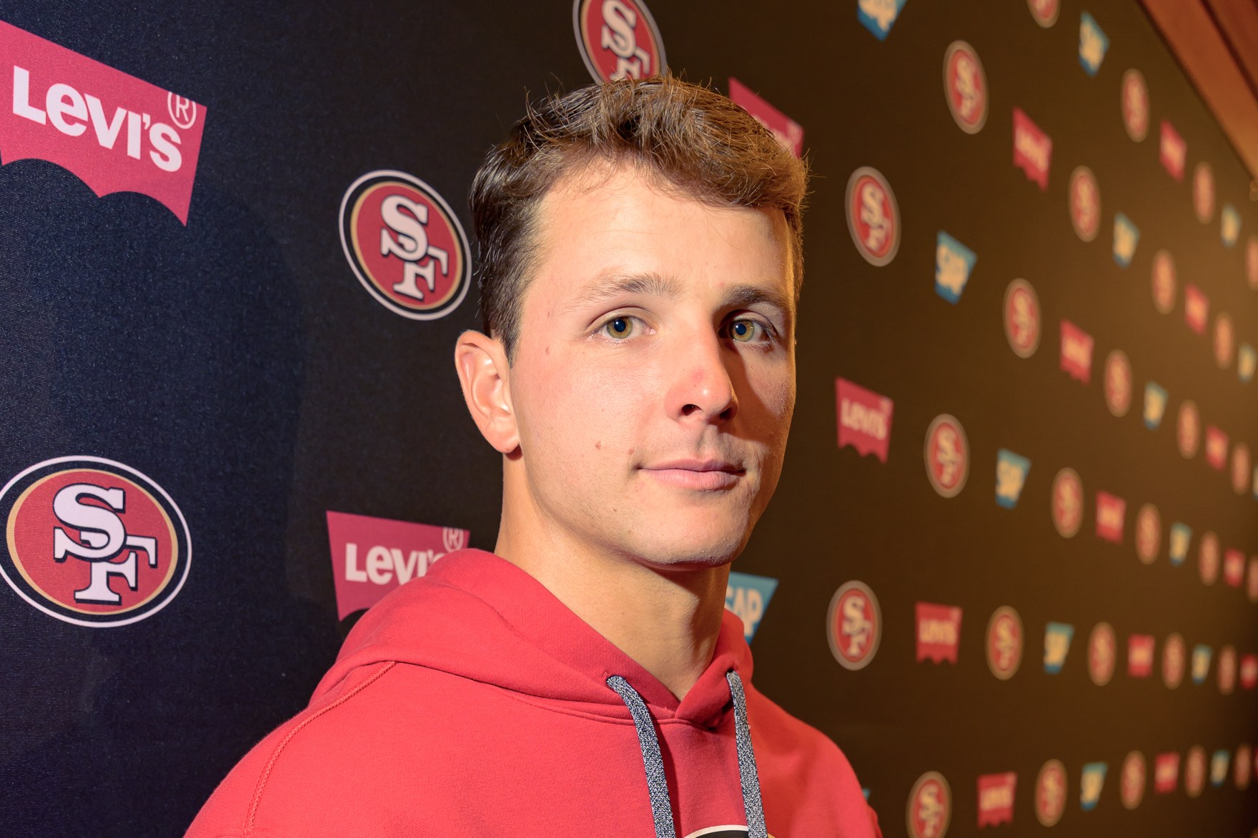 A person in a red hoodie with an "SF" logo stands against a backdrop featuring repeated logos of Levi's, SAP, and the same "SF" design.