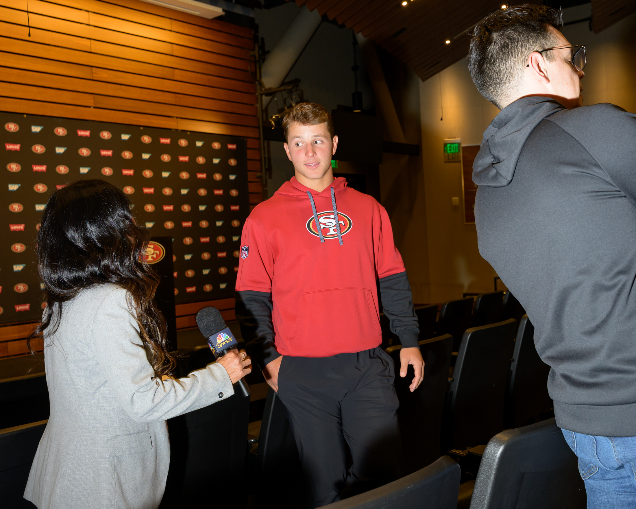 A young man in a red sports hoodie stands in a room facing a woman holding a microphone. A man in a black hoodie is partially visible on the right.