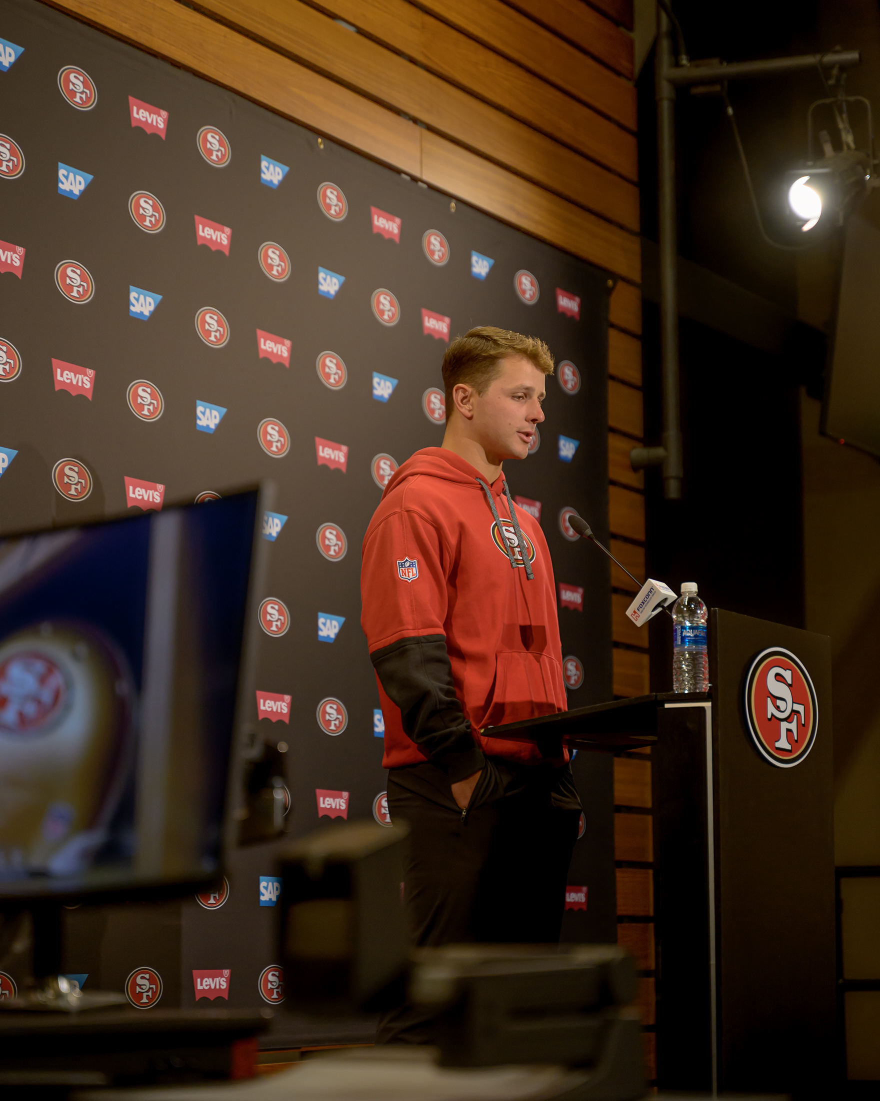 A person in a red 49ers hoodie stands at a podium with a microphone and water bottle. The backdrop features 49ers logos, SAP, and Levi’s branding.