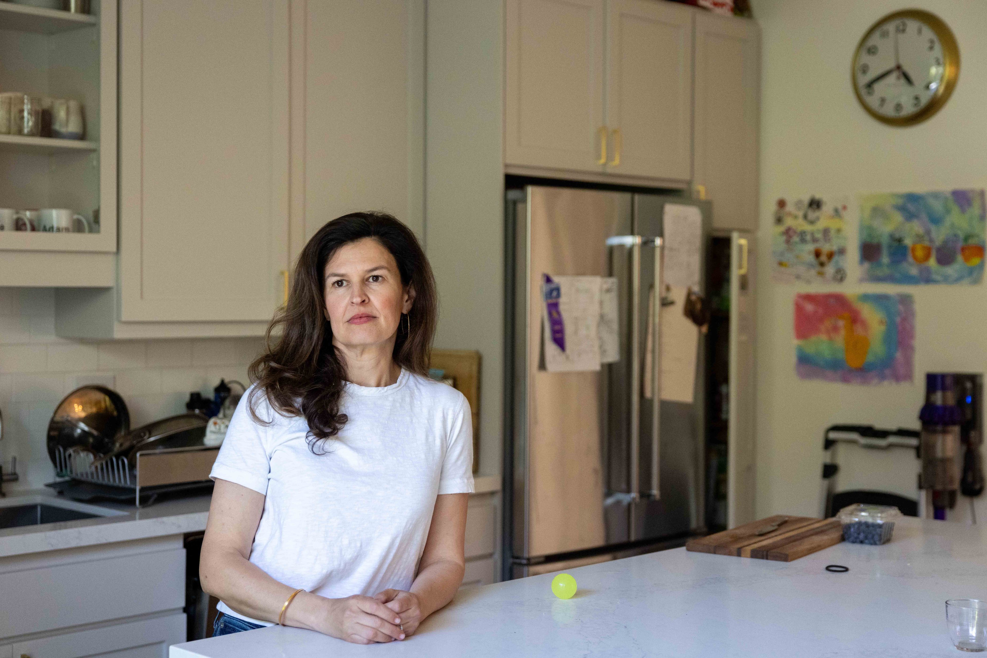 A woman in a white shirt stands in a kitchen with light cabinets and a refrigerator. A wall clock and children's drawings are visible in the background.