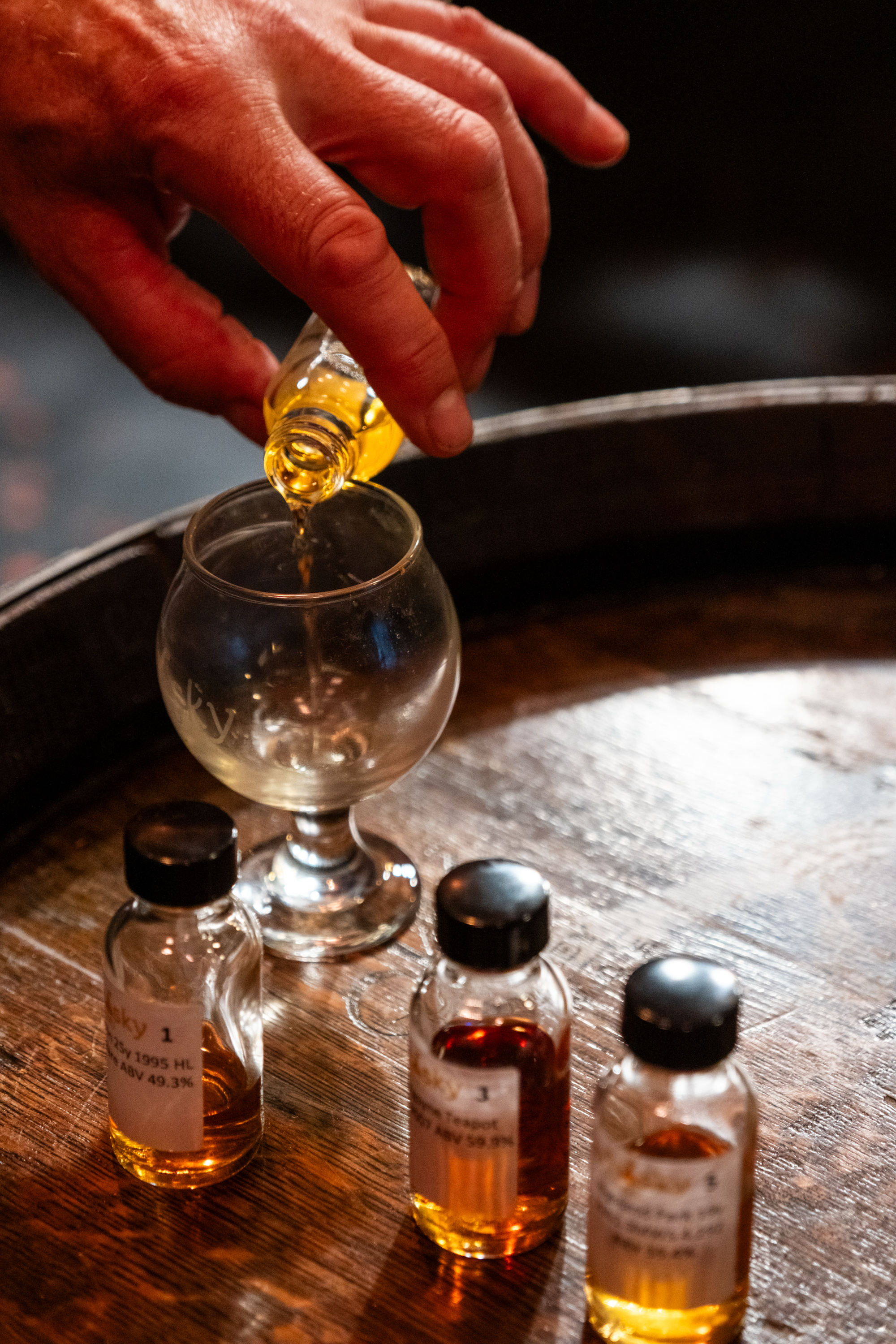 A hand pours a golden liquid from a small bottle into a glass on a wooden surface. Three small bottles with dark caps and labels are nearby.