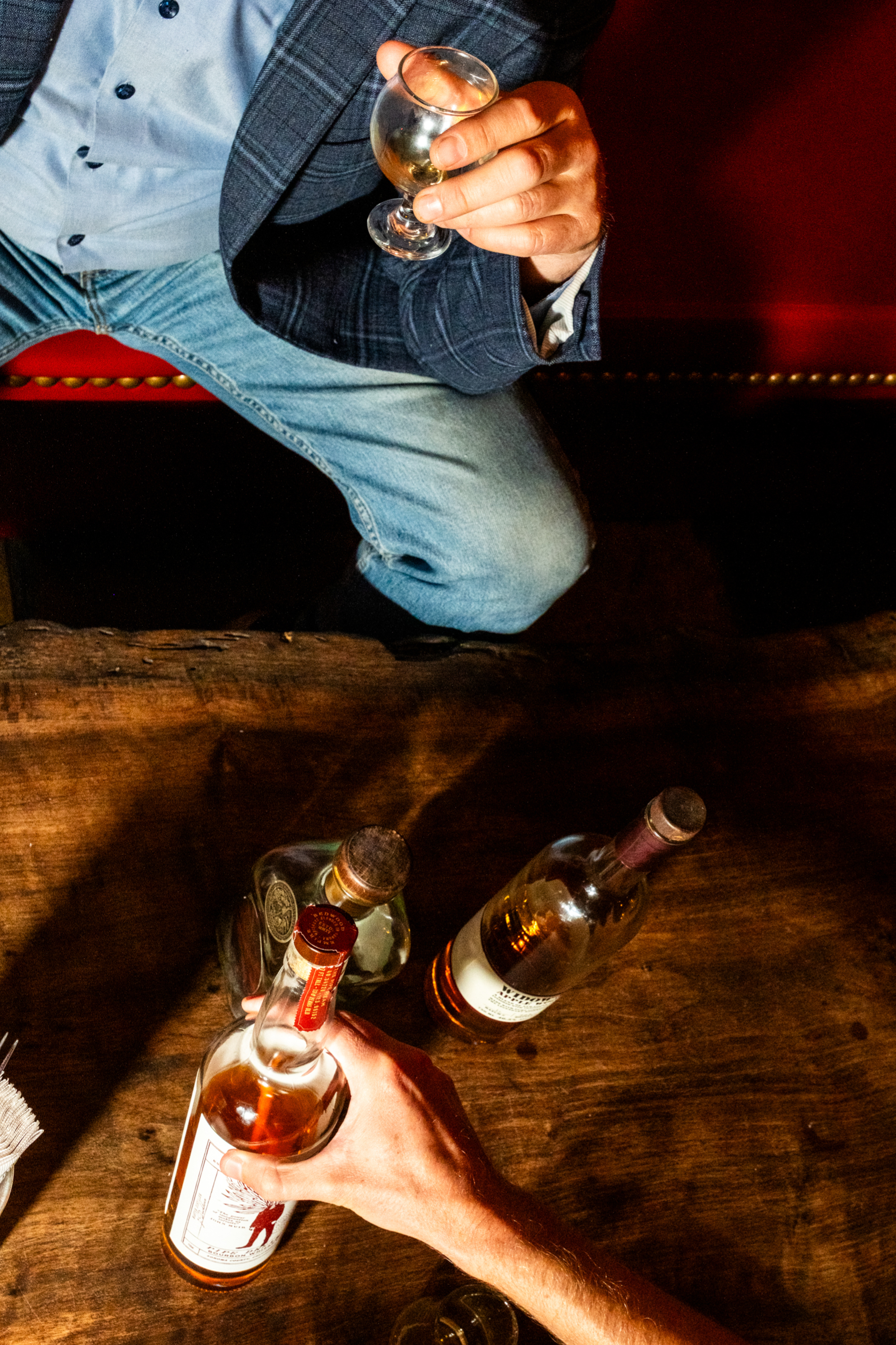 A person in a blue blazer and jeans holds a glass, seated at a wooden table with three bottles of alcohol, while another hand reaches for a bottle.