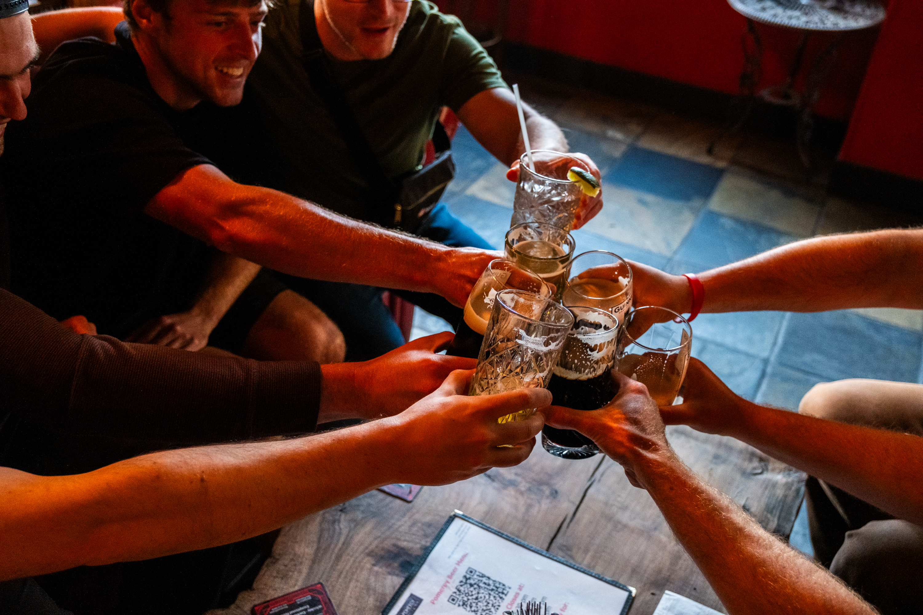 A group of people is holding various glasses, toasting together over a wooden table. Their arms are extended, and the atmosphere appears lively and celebratory.