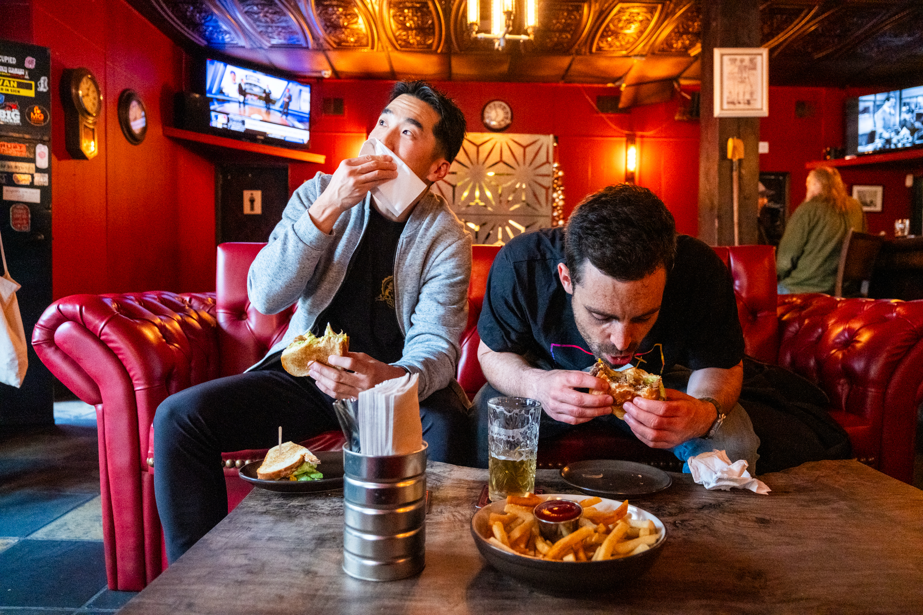 Two people sit on a red couch in a cozy, dimly-lit bar. They are eating burgers and fries, with one wiping his mouth and the other focusing intently on his meal.