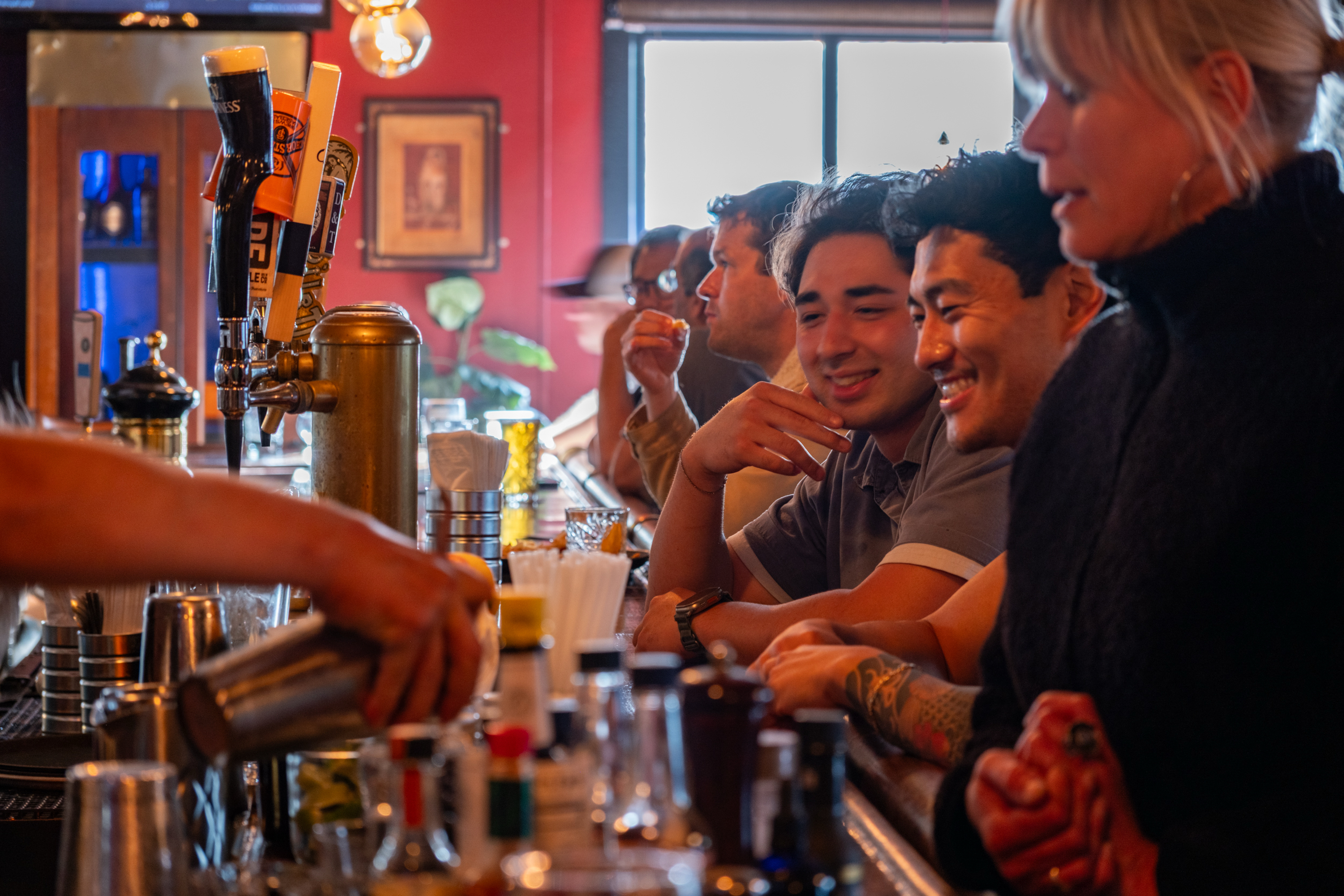 Several people sit at a bar, smiling and chatting. A bartender is pouring a drink. There are beer taps and various bar items in the foreground.