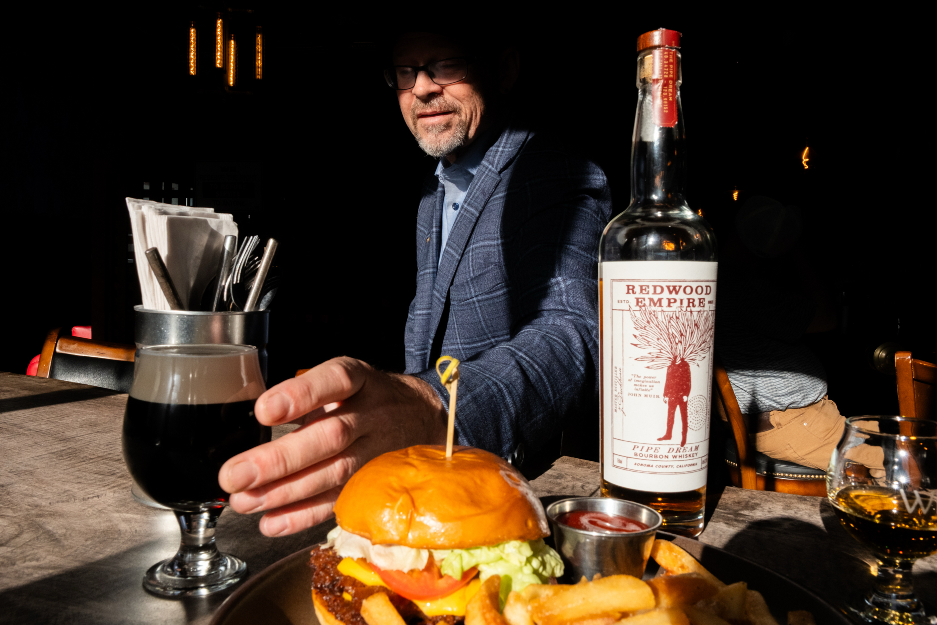The image shows a man sitting at a table with a burger and fries in front of him. He reaches for a dark drink. A bottle labeled "Redwood Empire" is on the table.