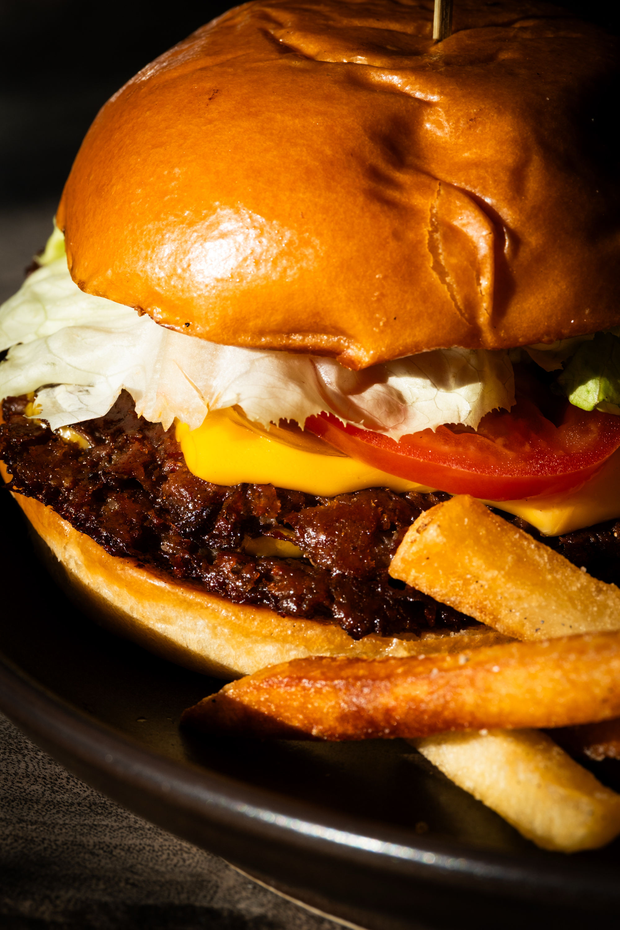 A close-up of a juicy burger with a shiny bun, lettuce, cheese, tomato, and a patty, served with crispy fries on a dark plate.