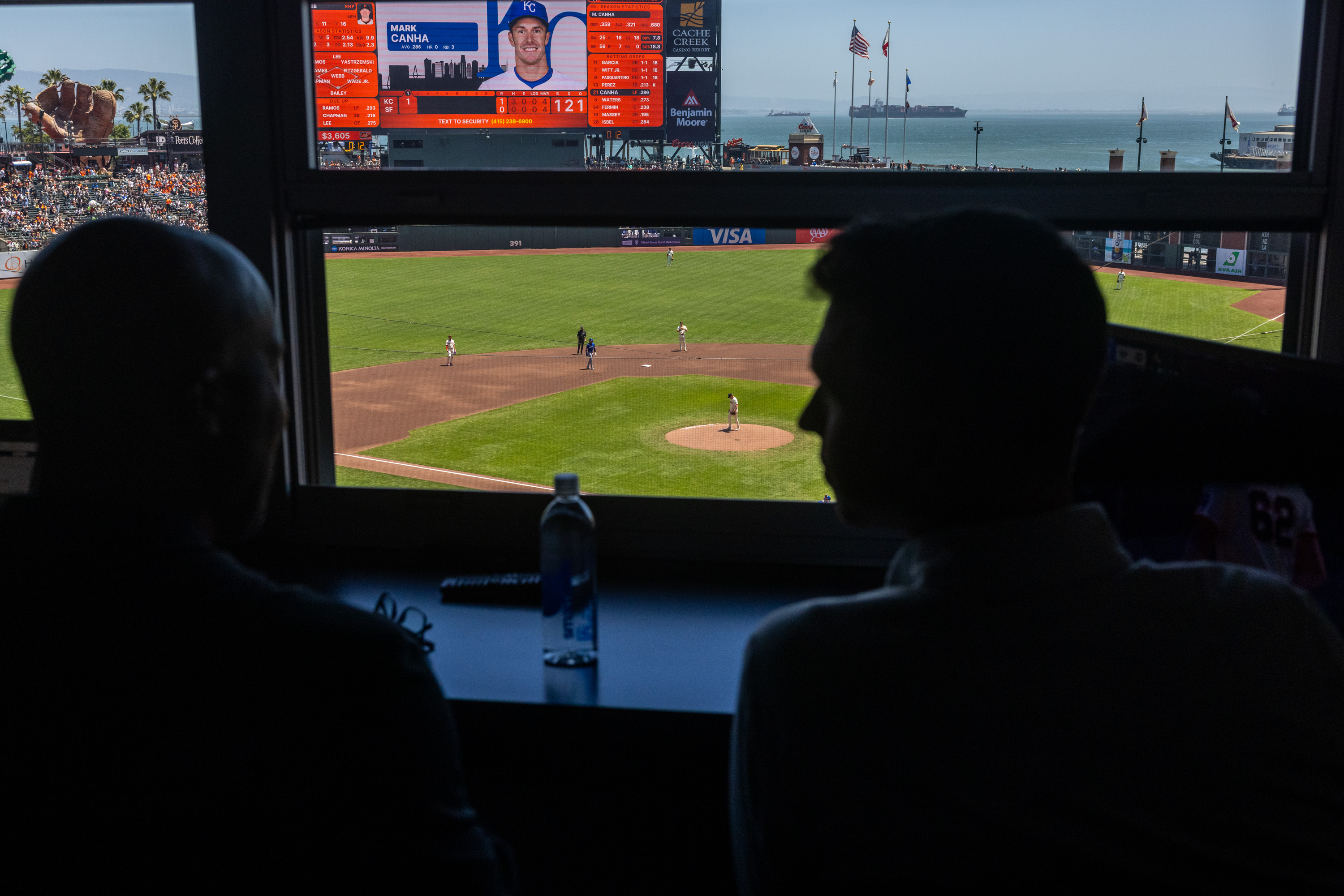 Two people in silhouette watch a live baseball game from a booth. The field is visible, with players positioned, and a player's profile appears on a screen above.