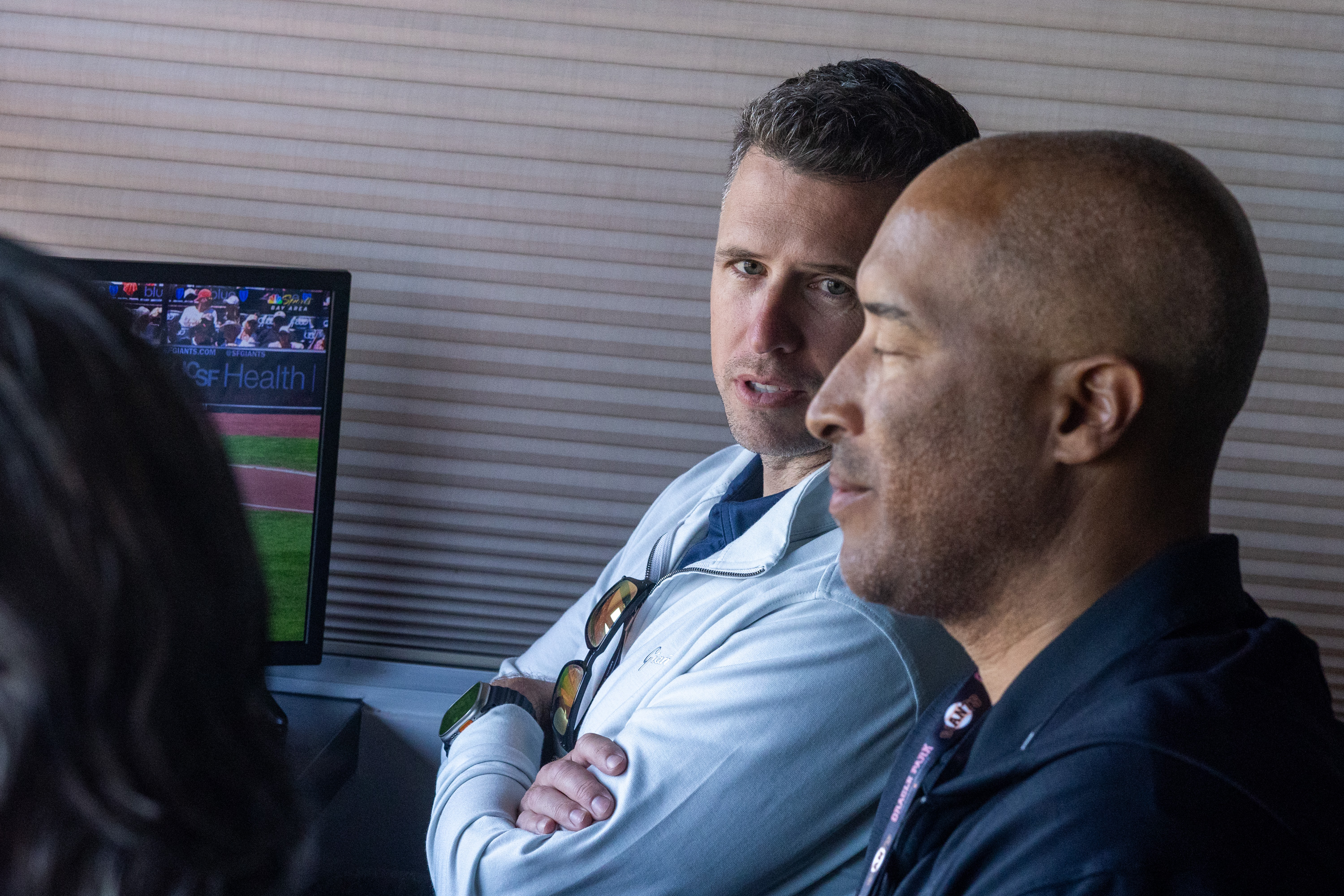 Two men are having a conversation in front of a monitor displaying a baseball game. One has short hair, wearing a light jacket with sunglasses on the collar.