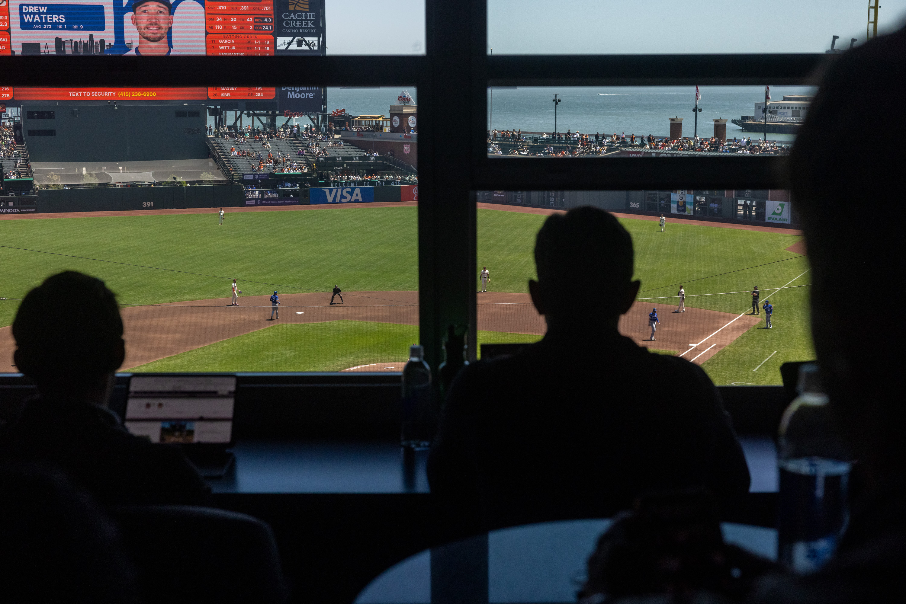 People watch a baseball game from inside a dark room with large windows. The diamond is visible with players on the field, and a big screen shows player stats.