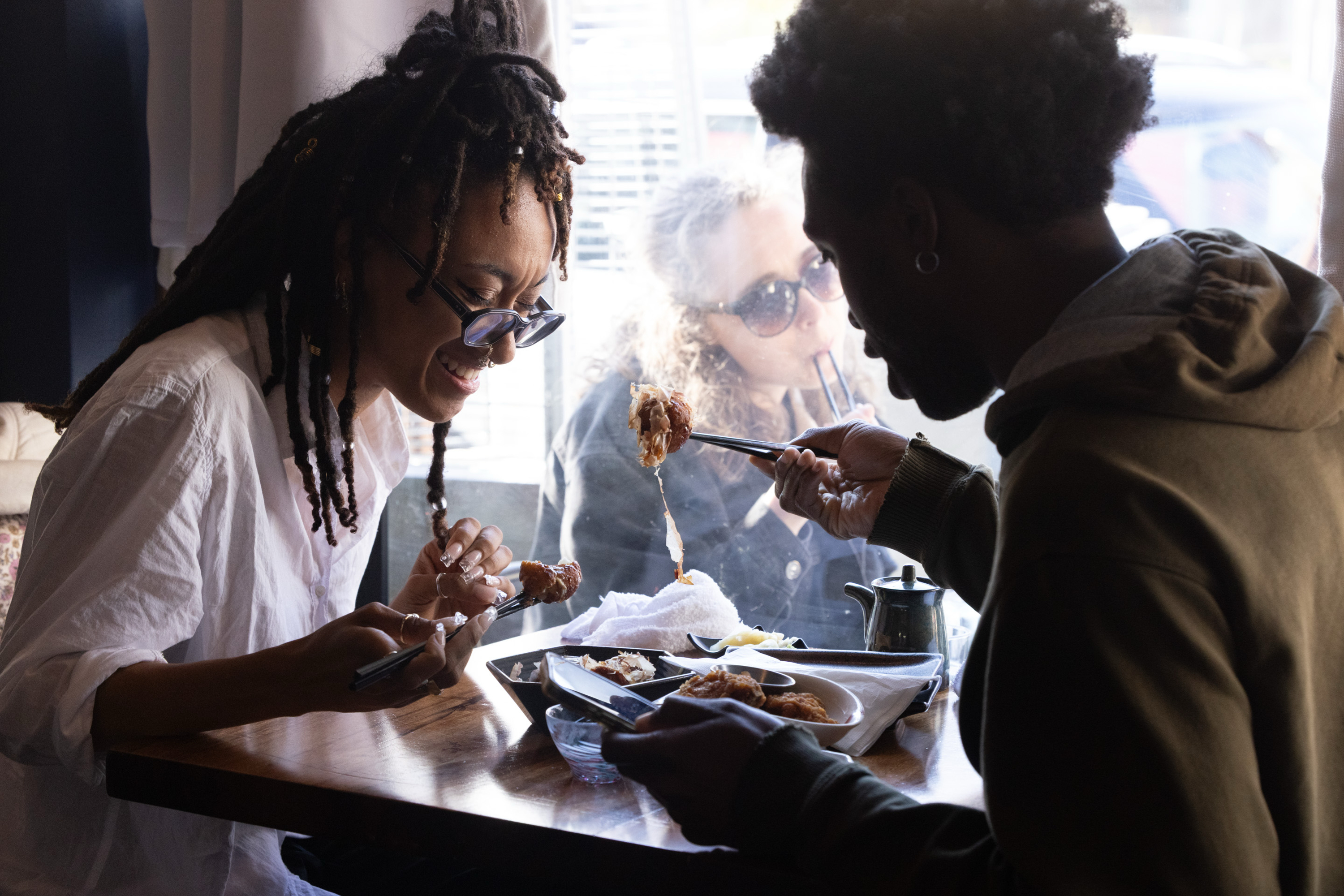 Two people are joyfully eating food at a wooden table, using chopsticks. A third person is visible through the window, sipping a drink with a straw.