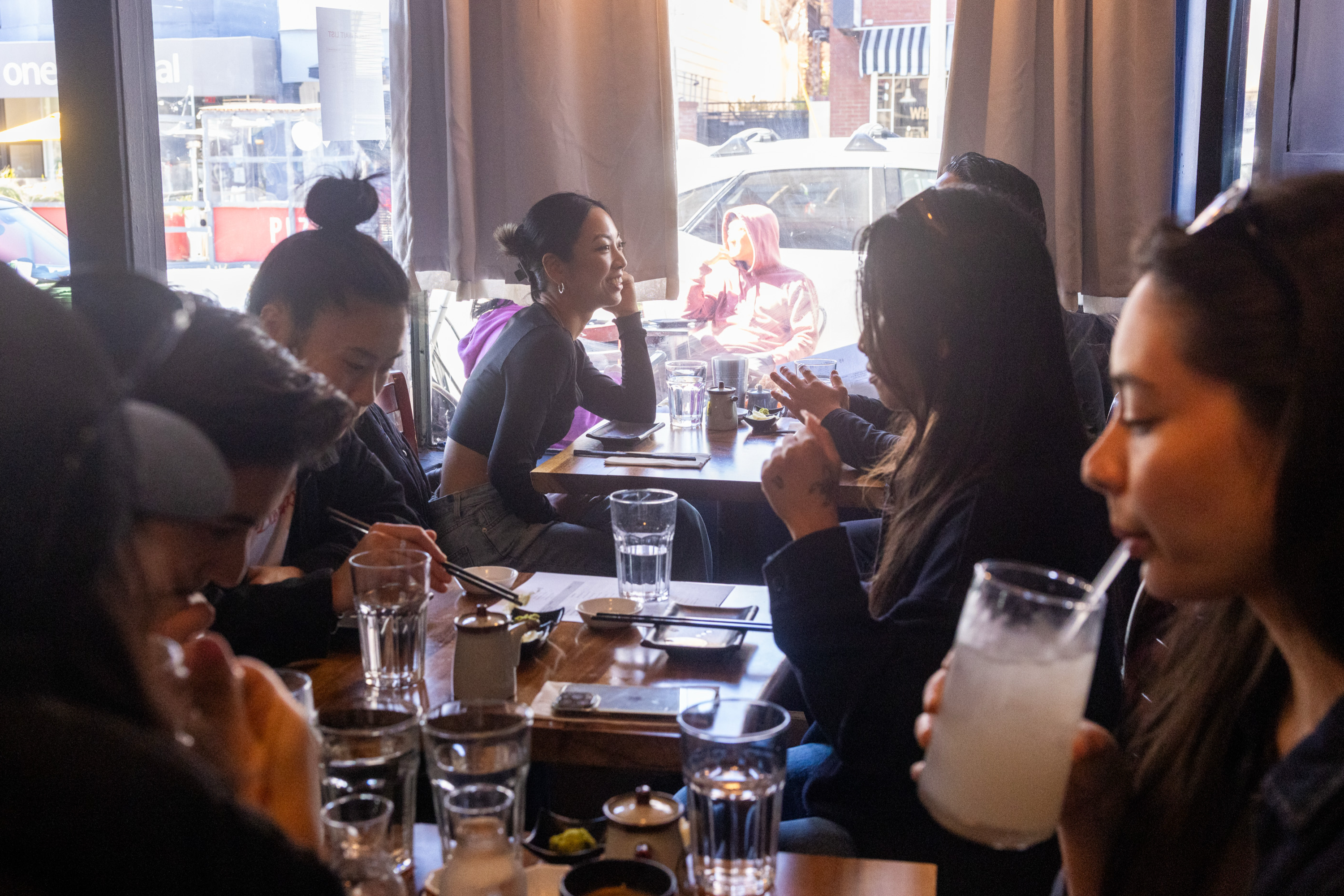 A group of people sit around a restaurant table, engaged in conversation. Glasses and small plates are on the table, and sunlight illuminates the scene.