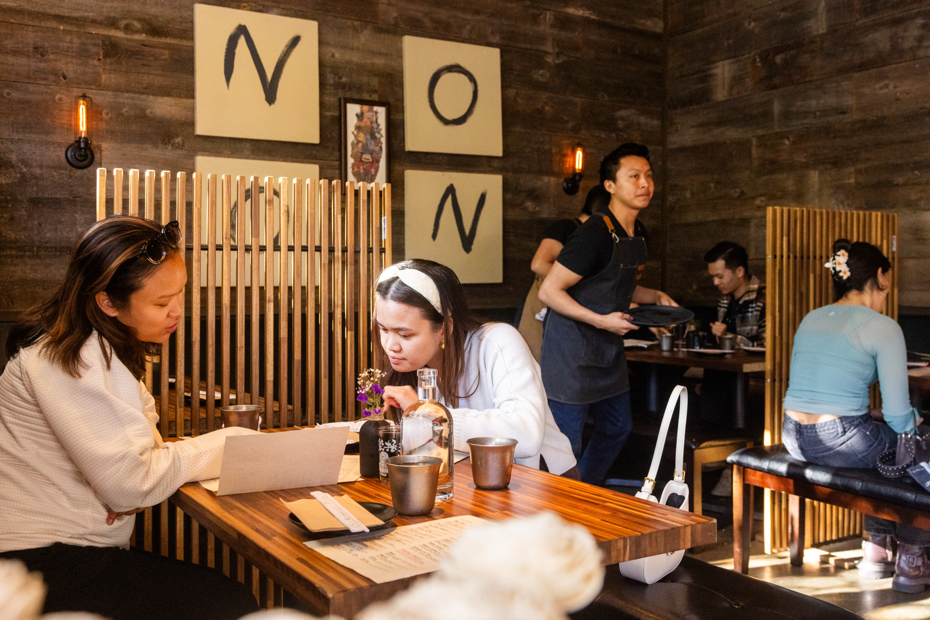 People are dining in a cozy restaurant with wooden accents. A server is carrying a tray, and two guests are focused on a menu at a wooden table.