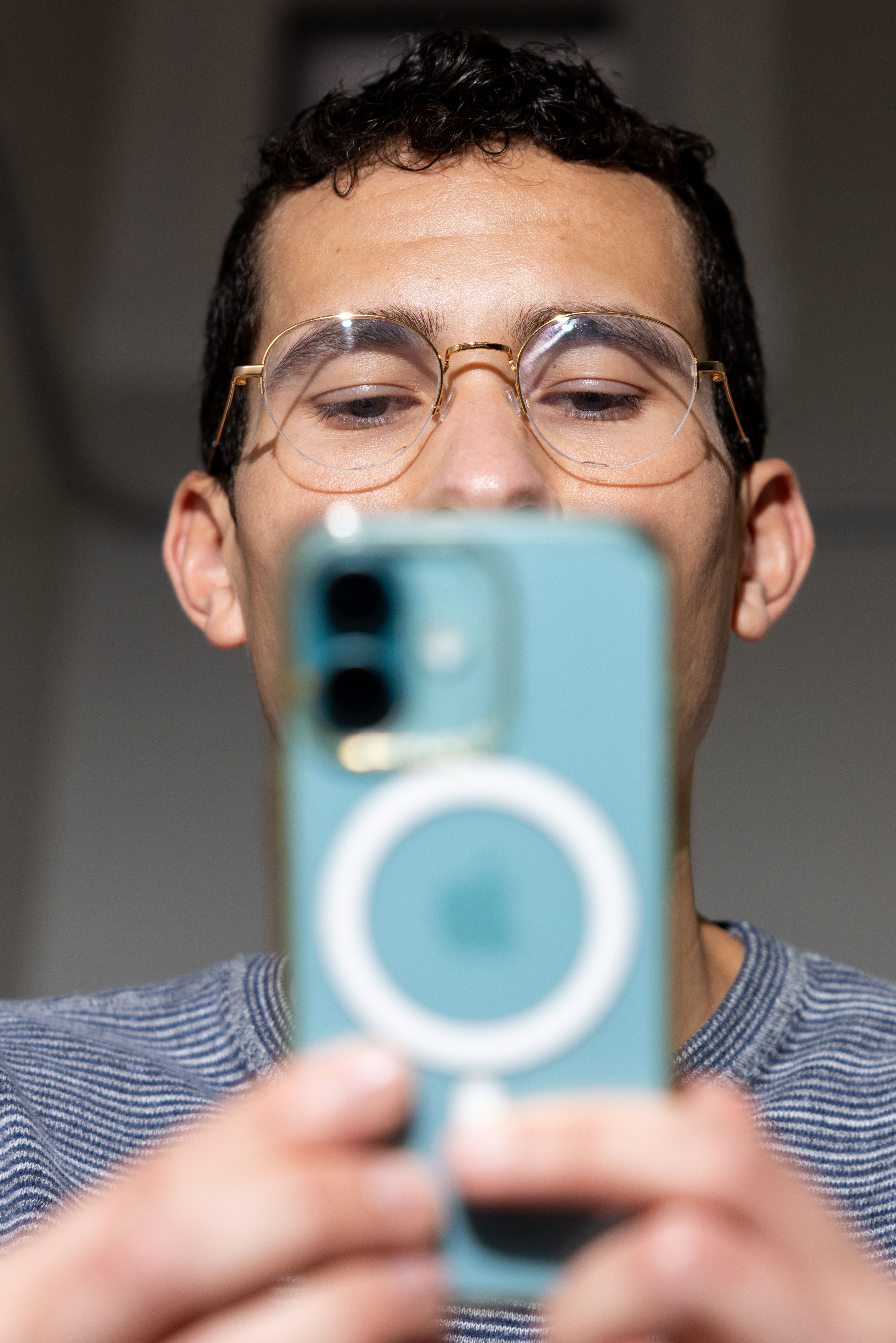 A person wearing glasses is taking a selfie with a turquoise iPhone. The focus is on the phone, with their face visible above it.