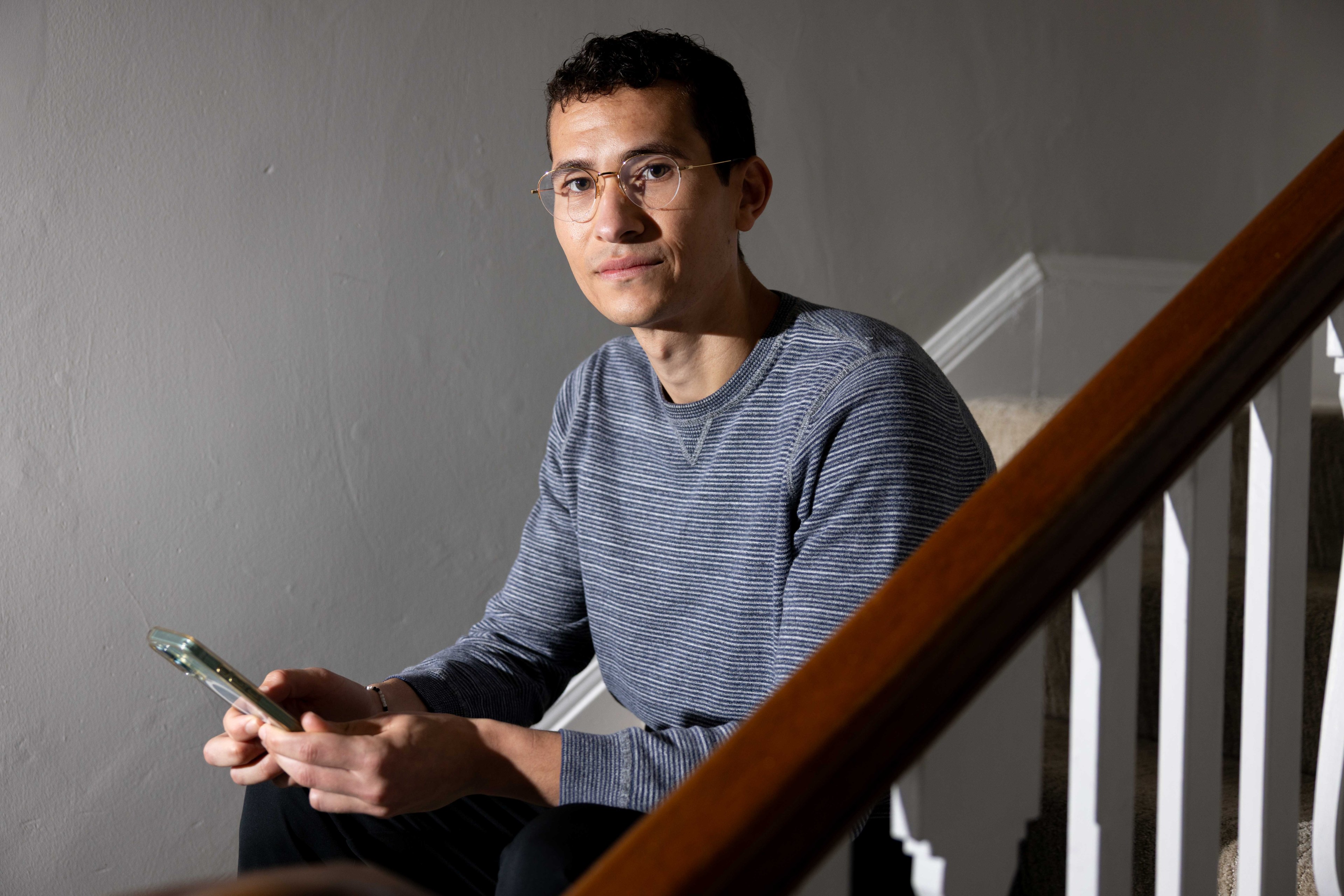 A person wearing a striped sweater and glasses sits on a staircase, holding a smartphone, against a plain gray wall.