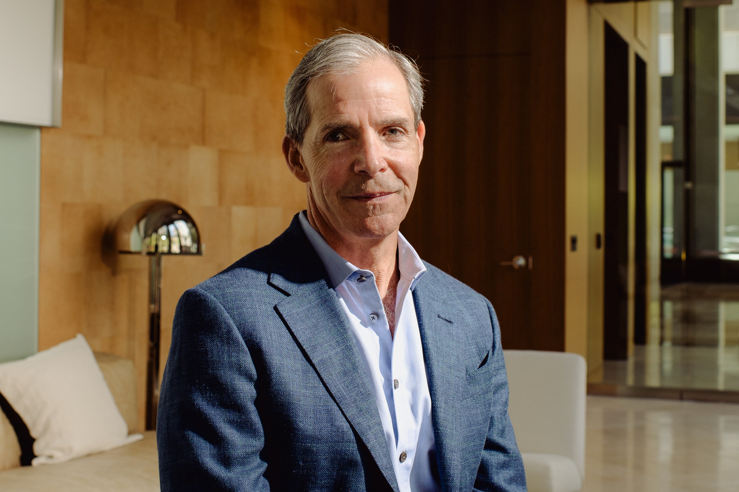 A man in a blue suit jacket and white shirt sits in a modern, warmly lit room with a sofa, lamp, and wooden paneled wall behind him.