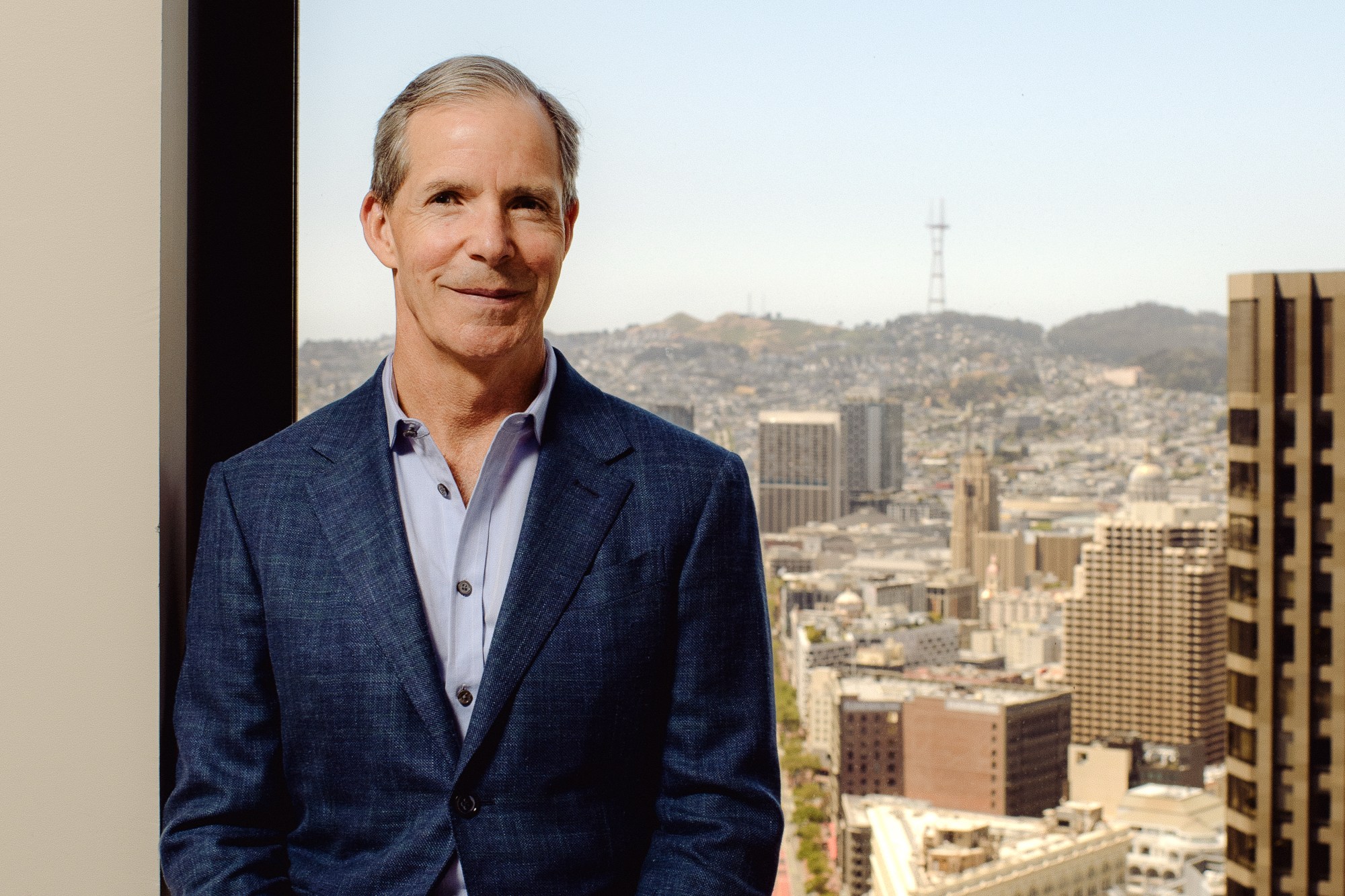 A man in a blue suit stands casually, leaning against a window ledge. Behind him is a vibrant cityscape with buildings and hills under a clear blue sky.