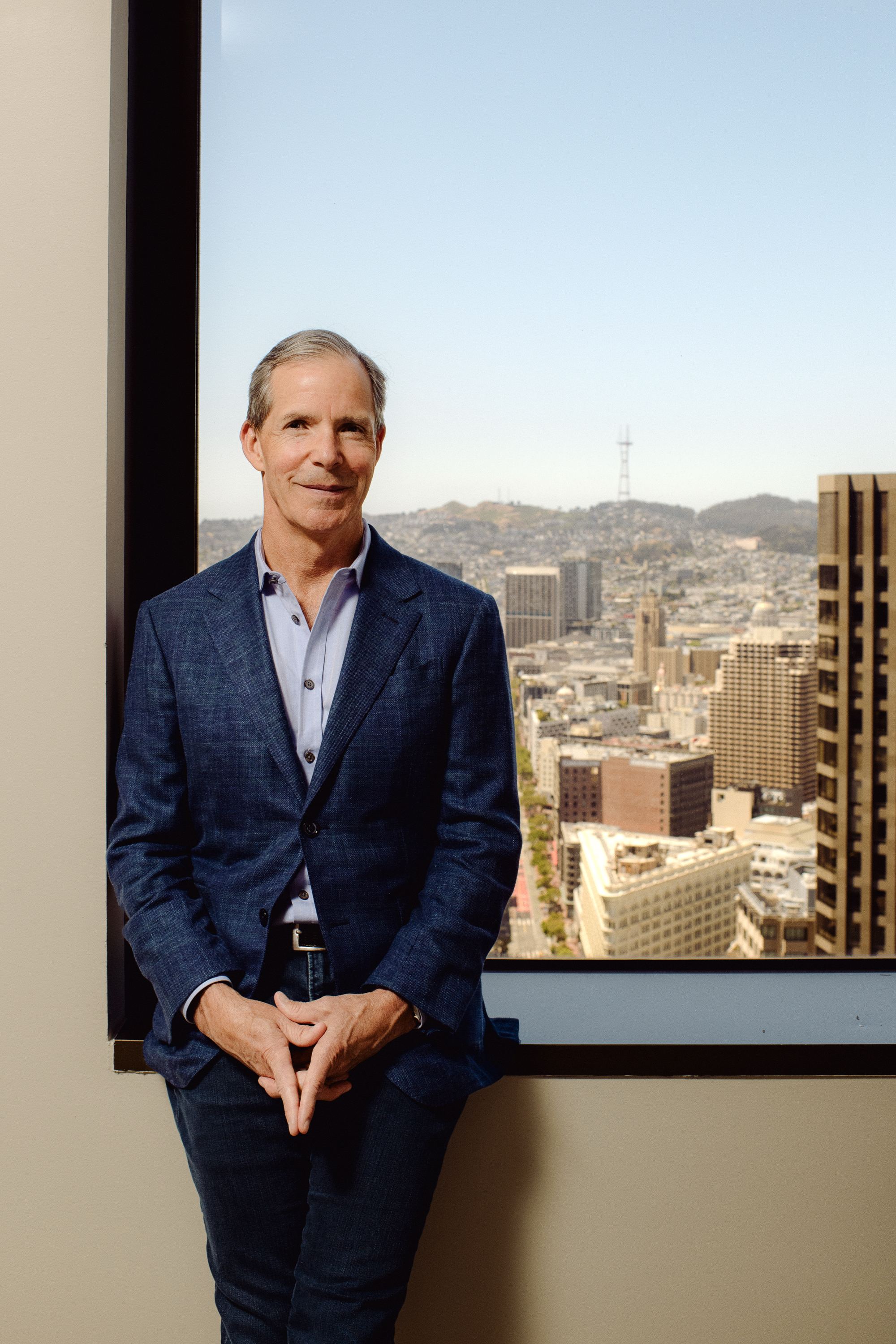A man in a blue suit stands casually, leaning against a window ledge. Behind him is a vibrant cityscape with buildings and hills under a clear blue sky.