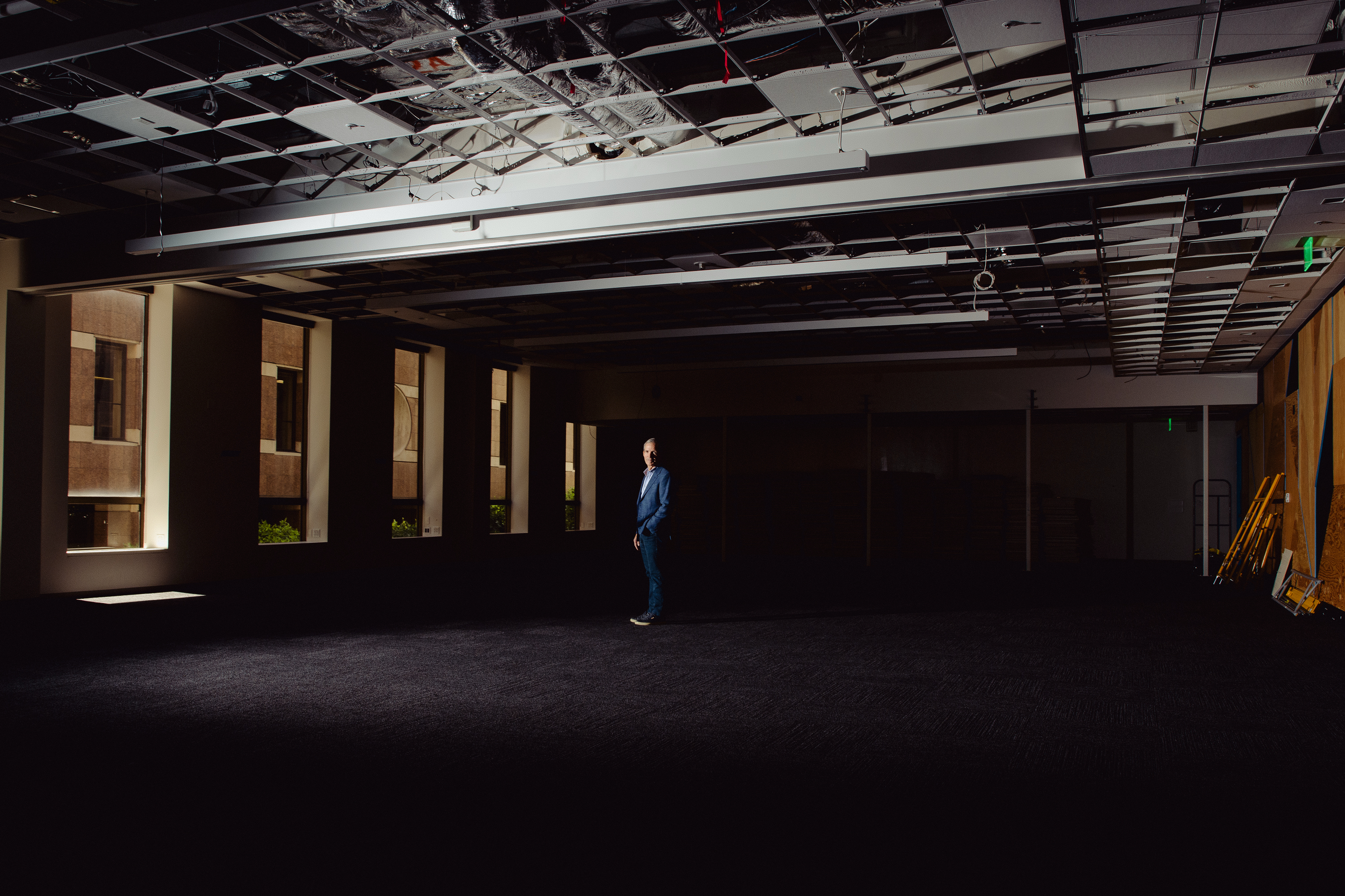A person in a blue suit stands in an empty, dimly lit room with exposed ceiling panels. Light filters in from windows on the left, casting shadows.