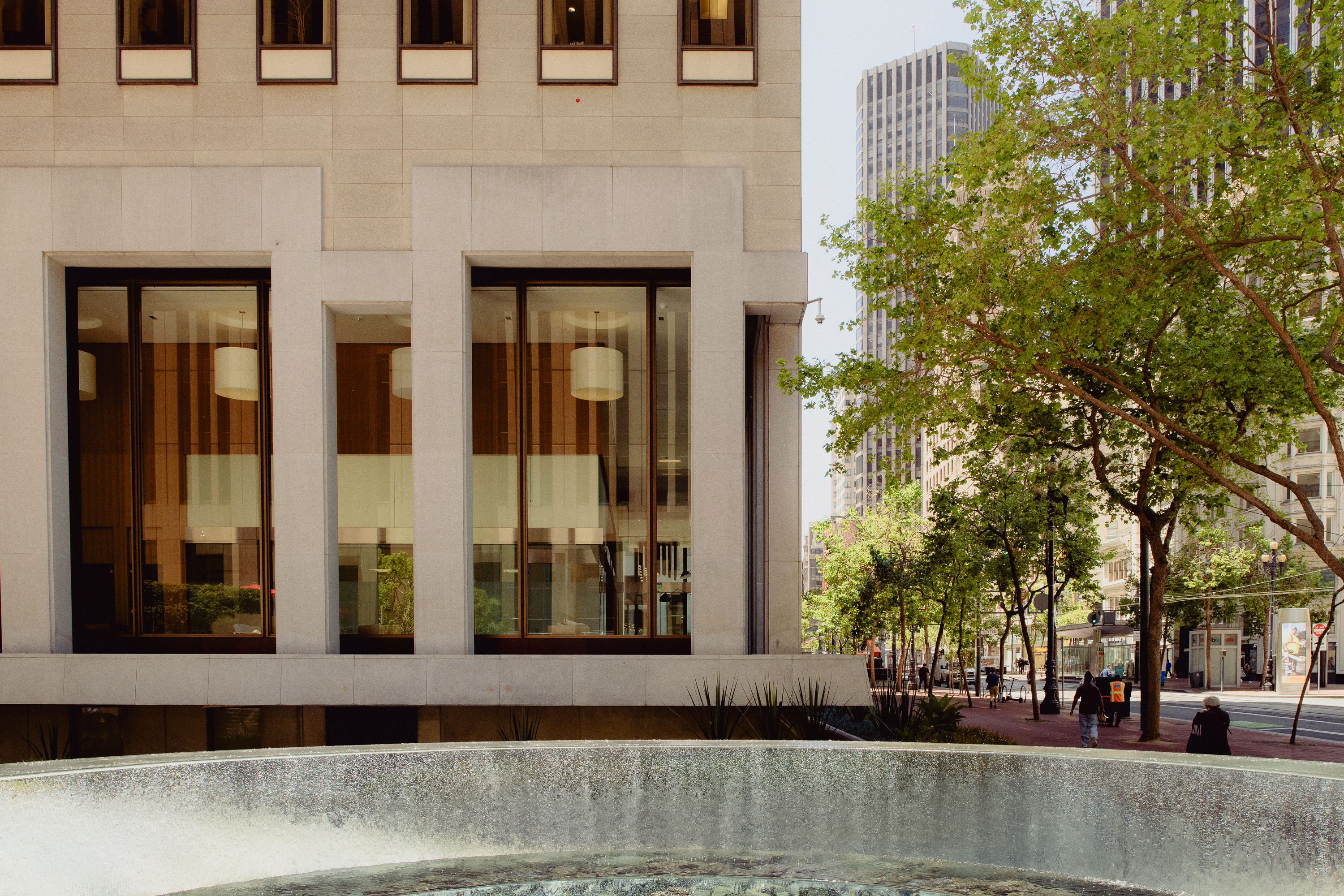 The image shows a modern building with large windows next to a tree-lined city street. In the foreground, there is a round water fountain.