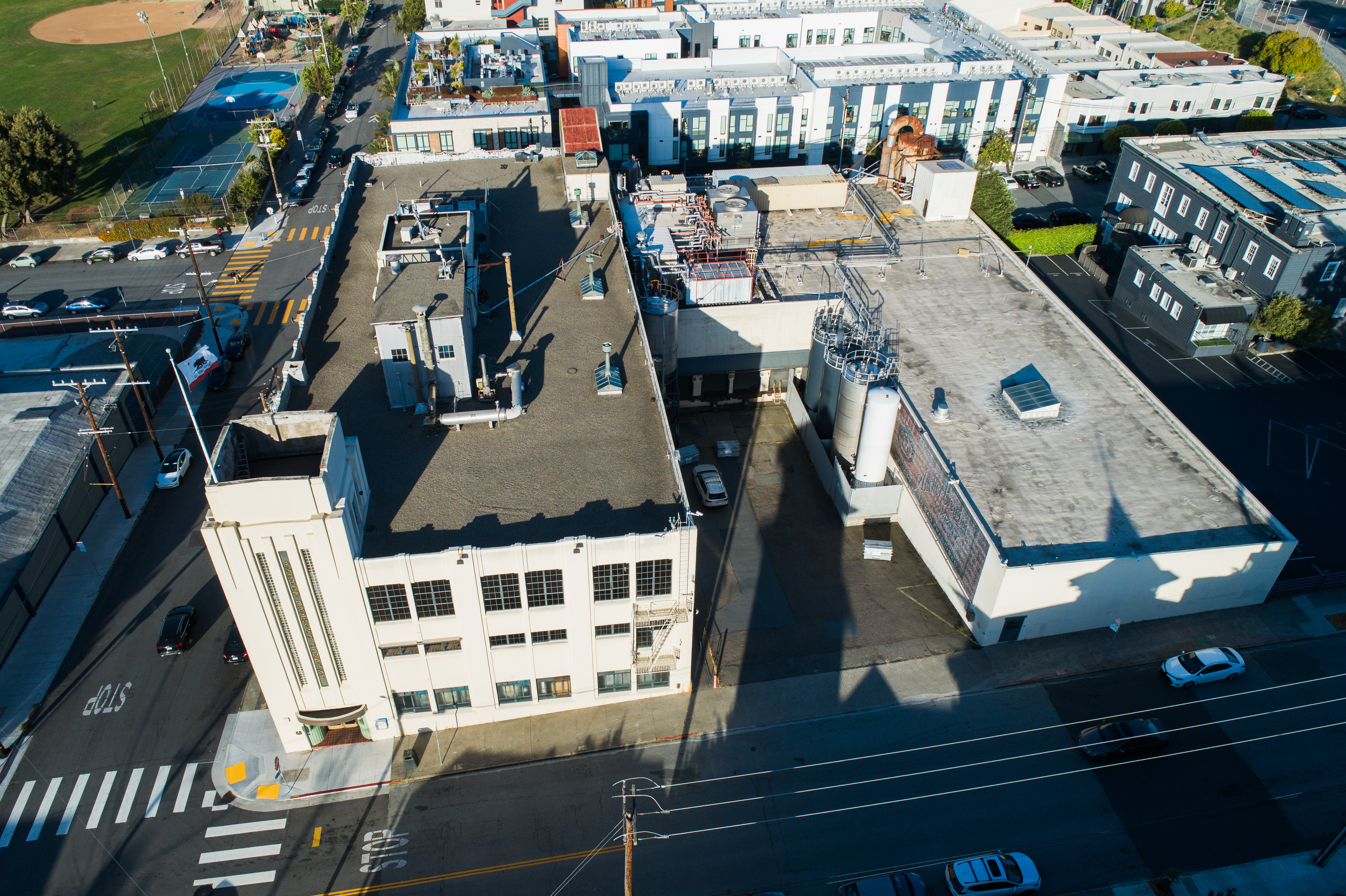 An aerial view shows an industrial building with a flat roof, surrounded by streets. Cars are parked nearby, and a sports field is visible in the background.