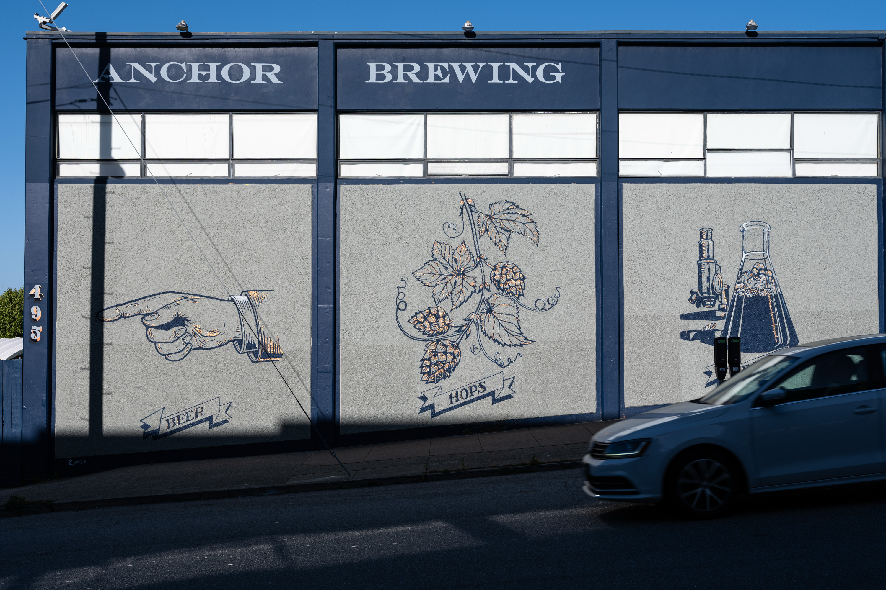 A wall mural with &quot;Anchor Brewing&quot; at the top. Illustrations show a pointing hand labeled &quot;Beer,&quot; hops, and a beaker. A car passes in front.