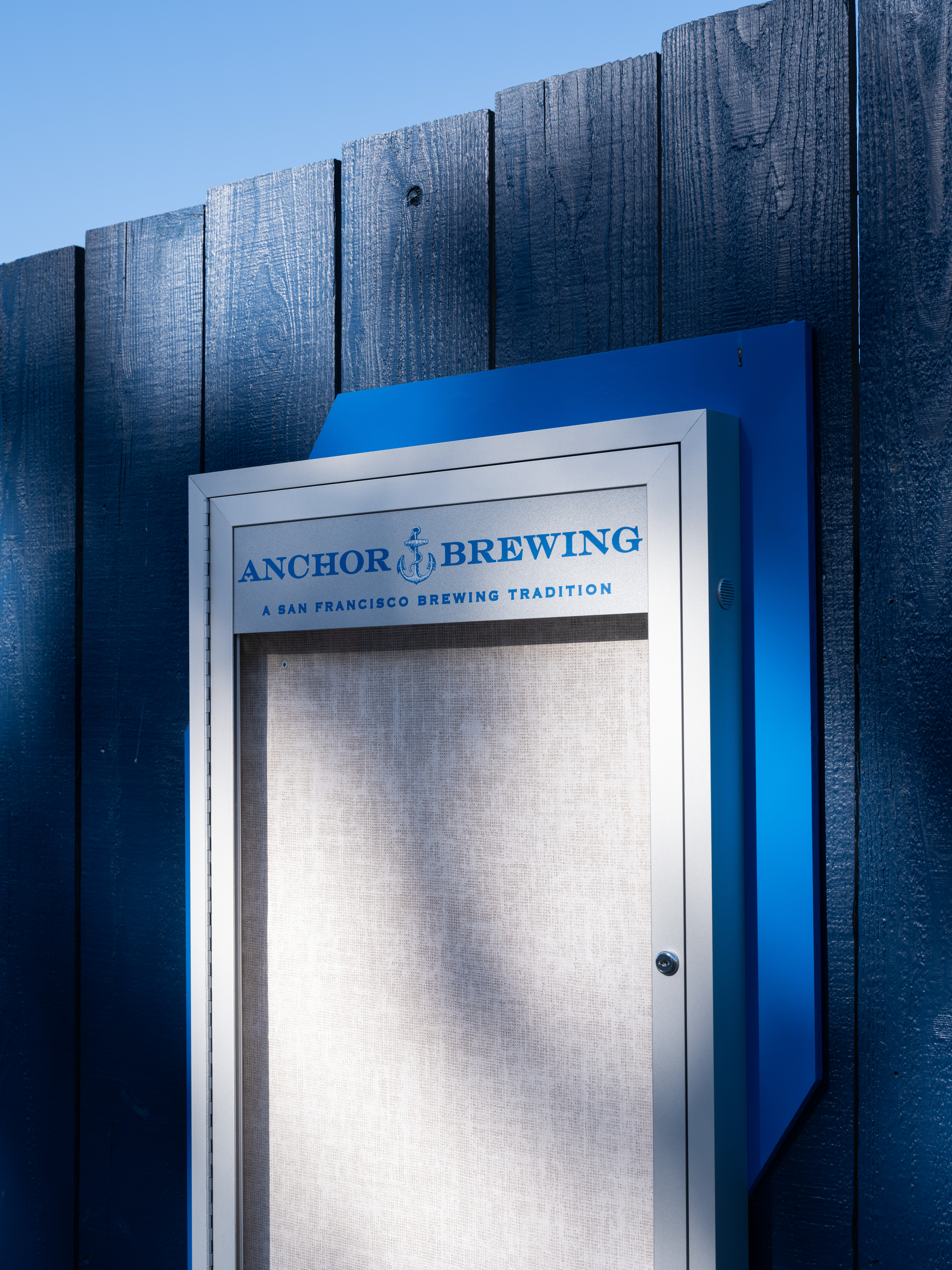 A blue and silver sign on a wooden fence reads &quot;Anchor Brewing: A San Francisco Brewing Tradition.&quot; Sunlight casts shadows across the empty board.