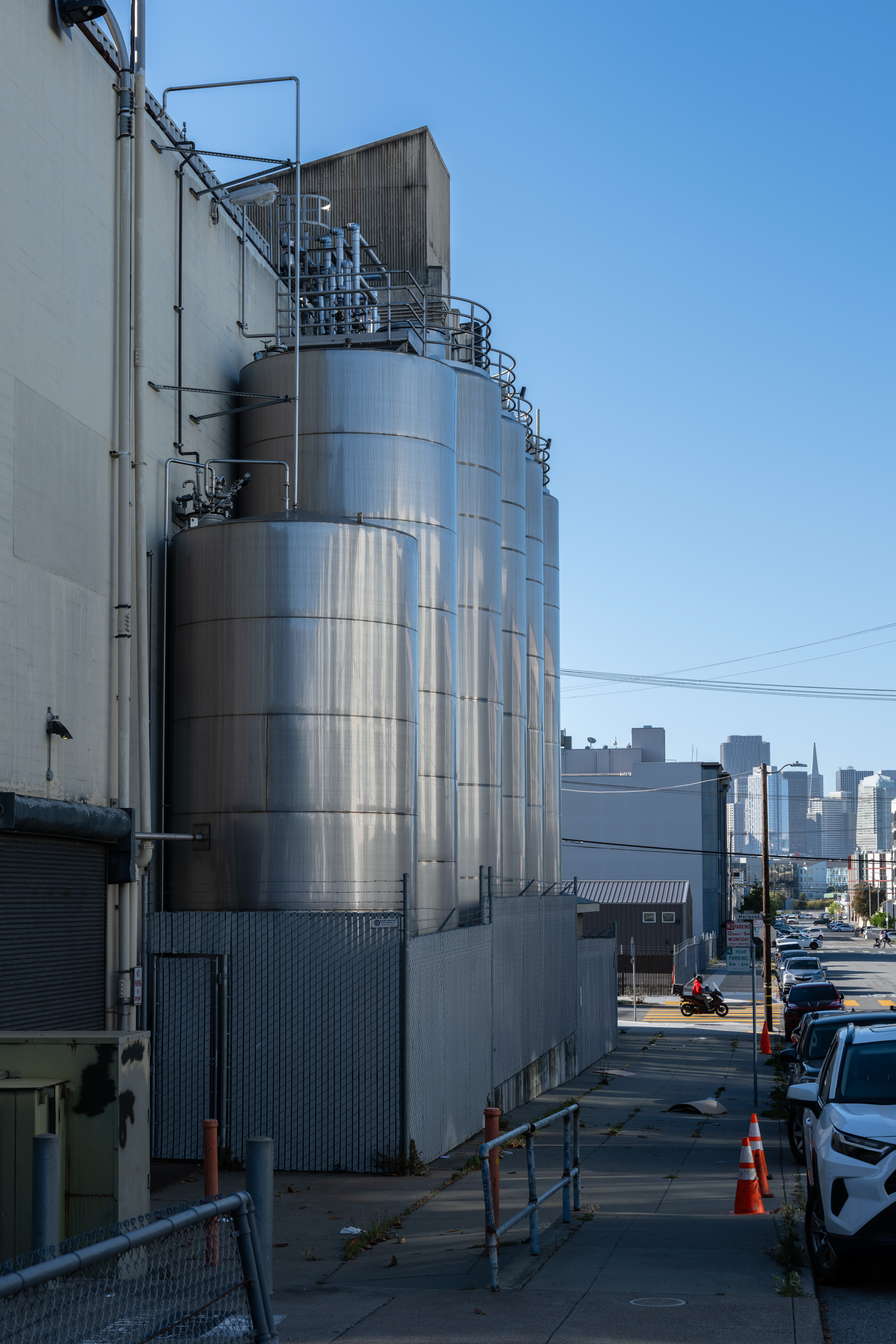 Large metal storage tanks are lined up along an industrial building. A street with cars and a distant city skyline are visible under a clear blue sky.