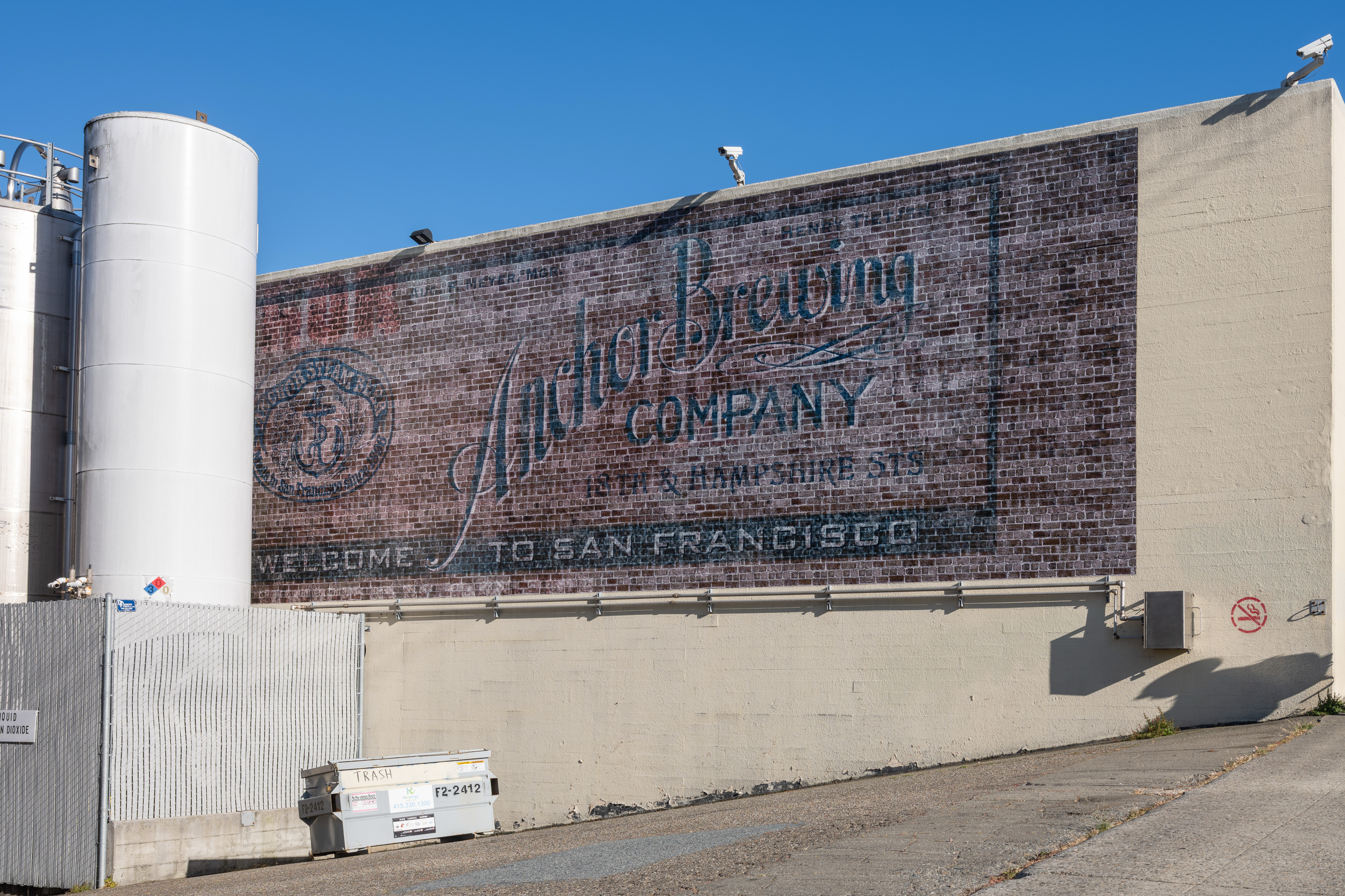 A faded mural on a brick wall reads &quot;Anchor Brewing Company&quot; with &quot;Welcome to San Francisco.&quot; Nearby are large white tanks and a small trash dumpster.