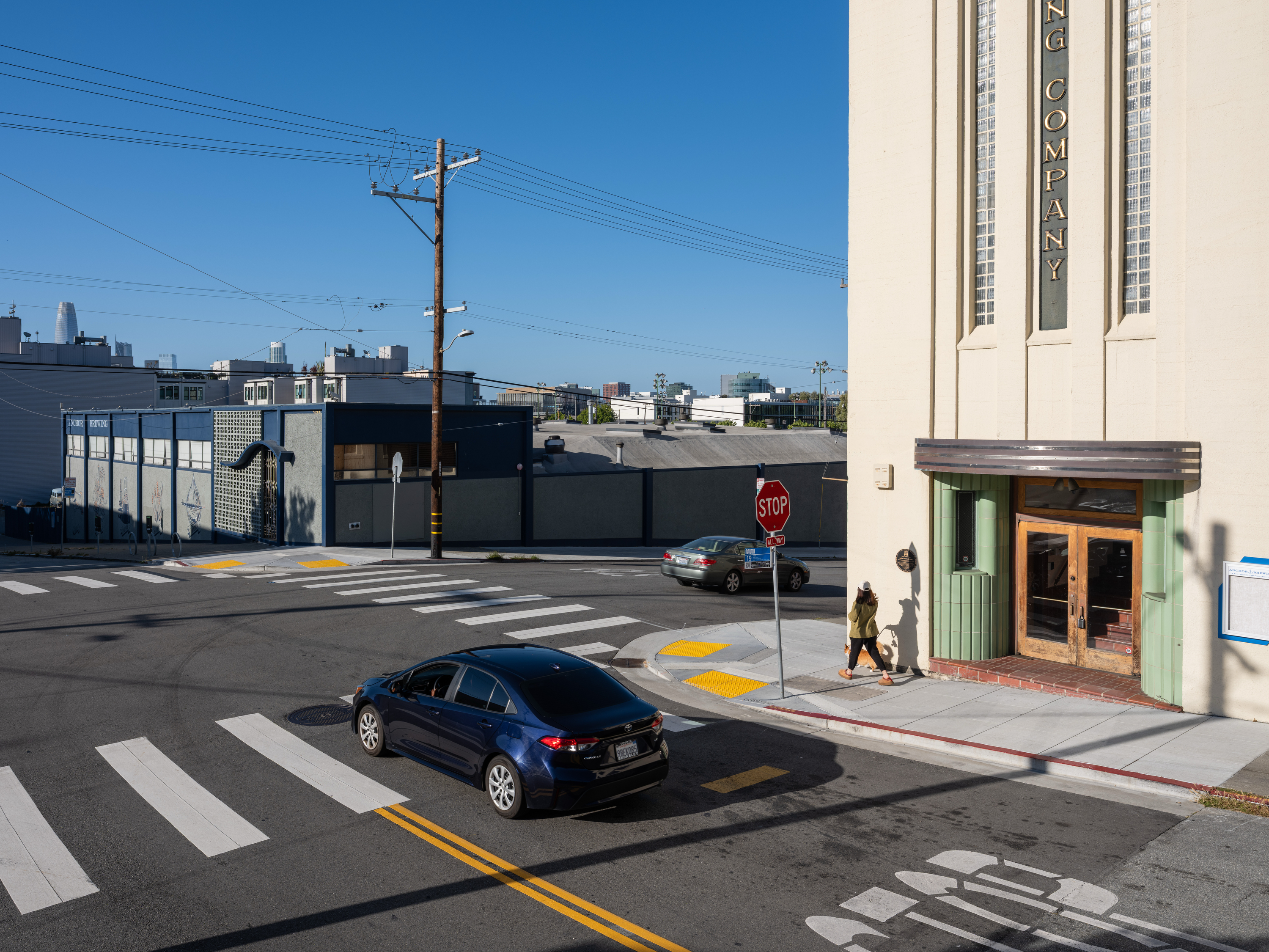 A person walks near a large beige building with &quot;COMPANY&quot; signage. A dark blue car waits at a stop sign on a sunny street with crosswalks.
