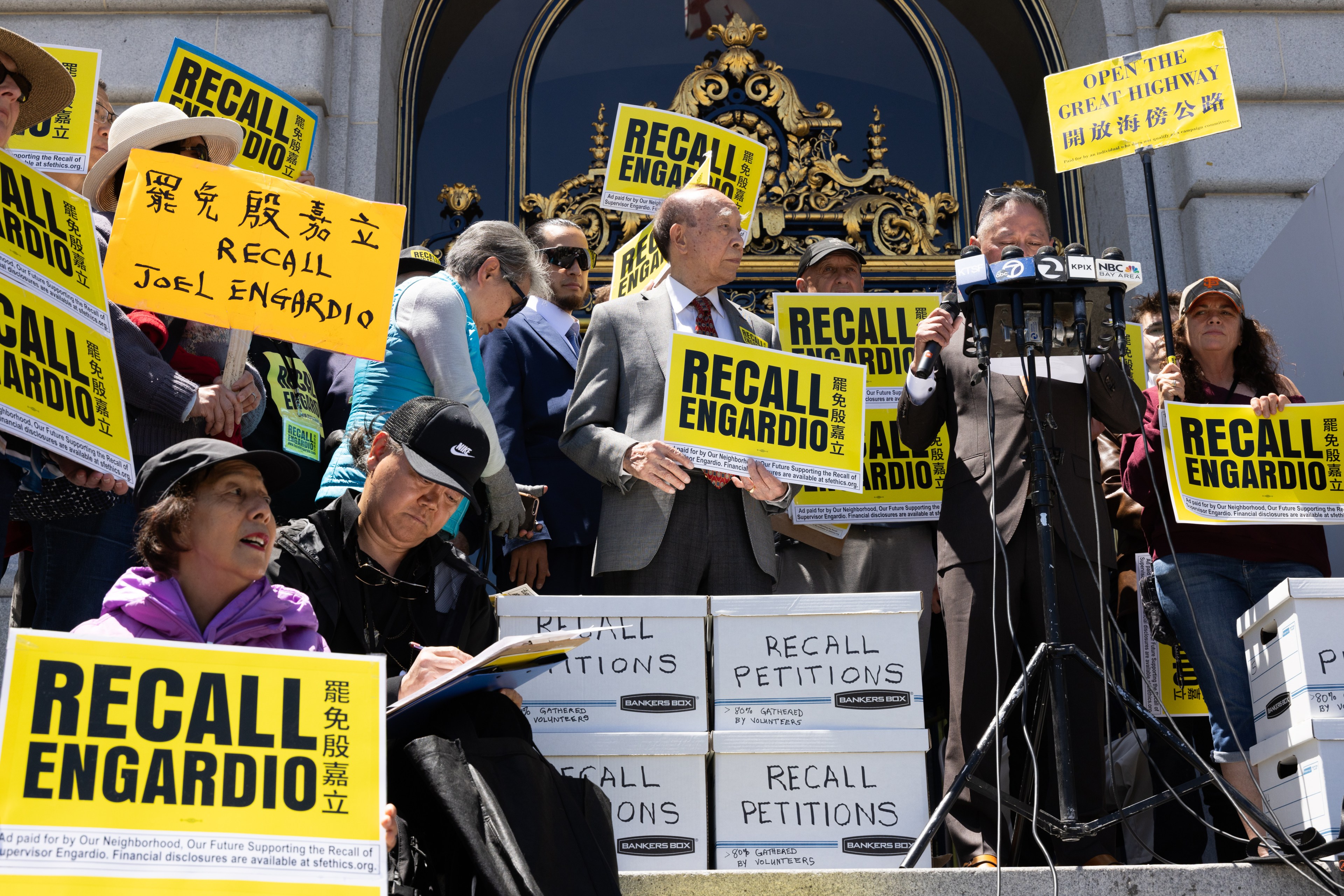 A group of people stands on steps holding &quot;Recall Engardio&quot; signs. Microphones and boxes labeled &quot;Recall Petitions&quot; are visible, indicating a public protest.