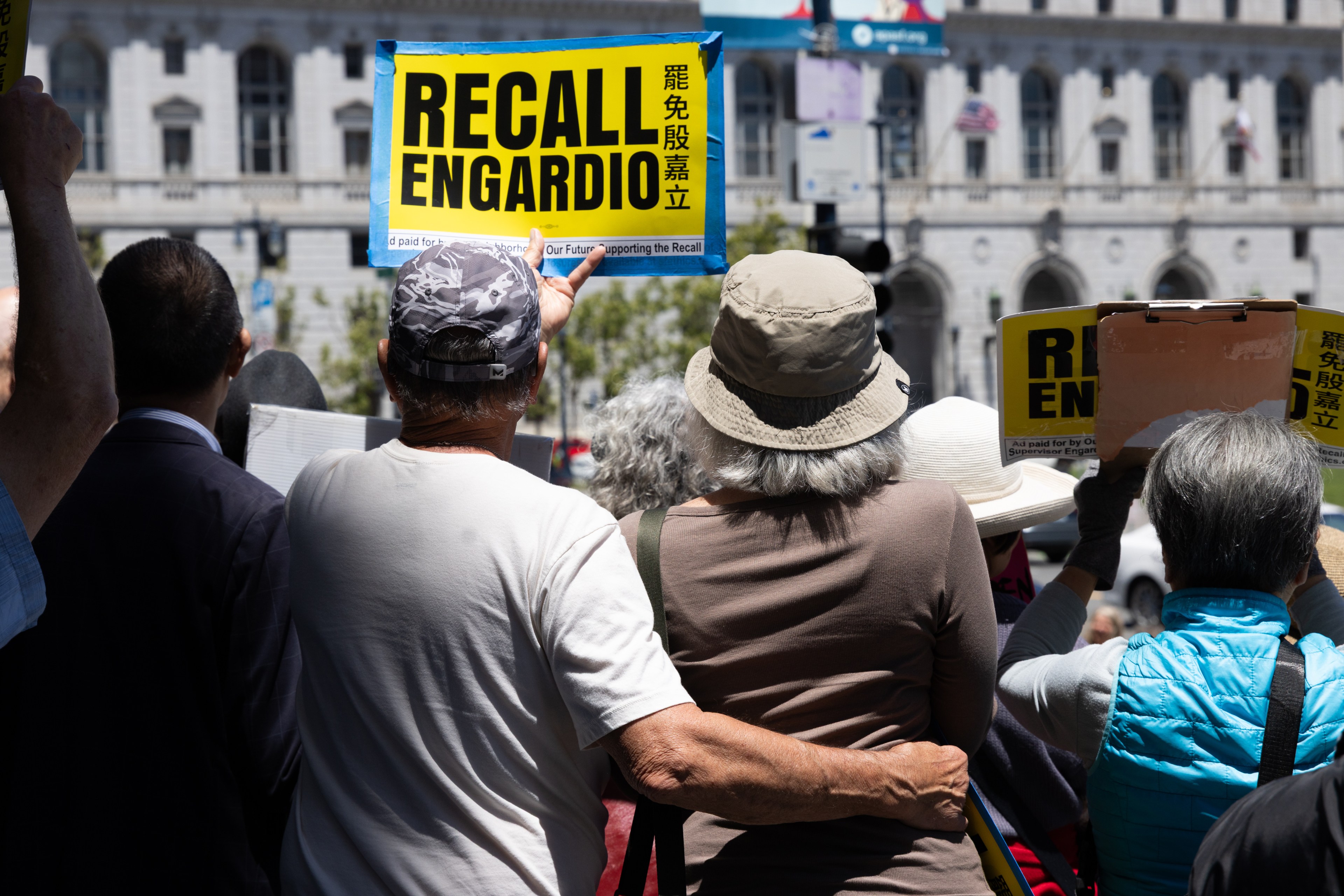 A small group of people stand closely together, holding signs that read "Recall Engardio" in English and another language, in an outdoor setting.