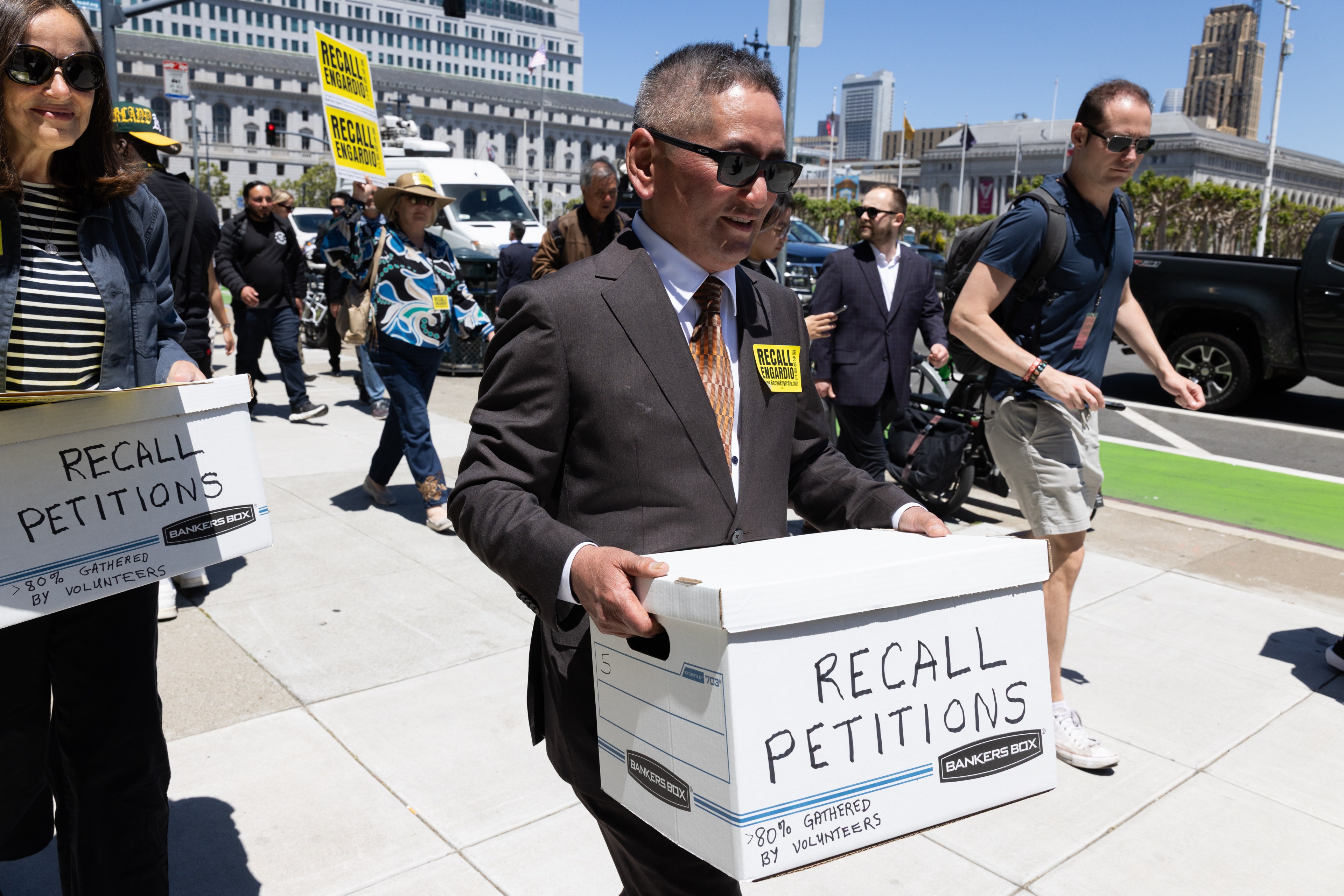 People are walking on a city street, carrying boxes labeled "Recall Petitions" and signs advocating recall. Some are in formal attire, and a large building is in the background.