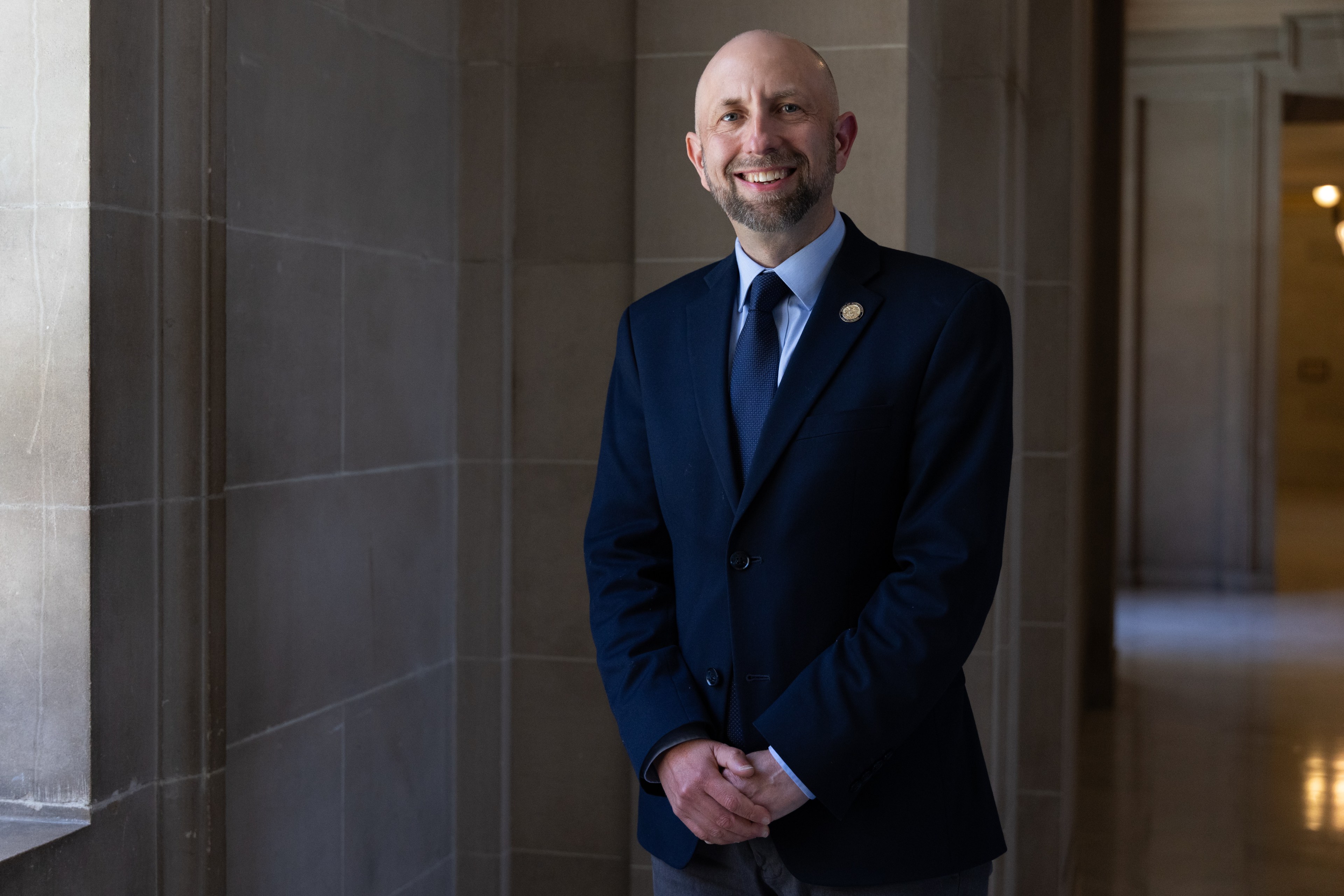 A man with a beard in a navy suit and tie is smiling while standing in a dimly lit corridor with gray stone walls.