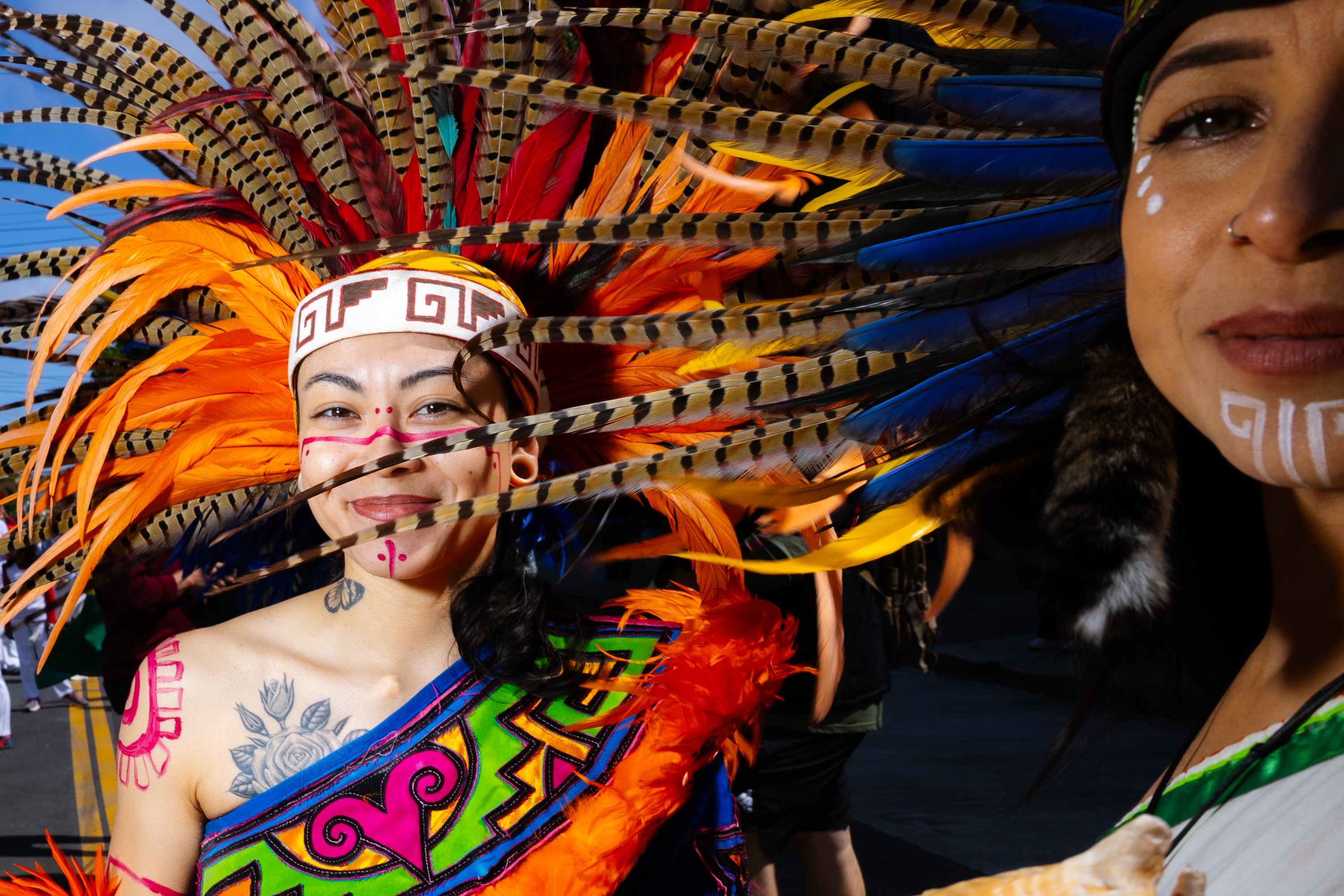 Two people in vibrant traditional attire, adorned with colorful feathers and face paint. One holds a conch shell. The background is a clear blue sky.