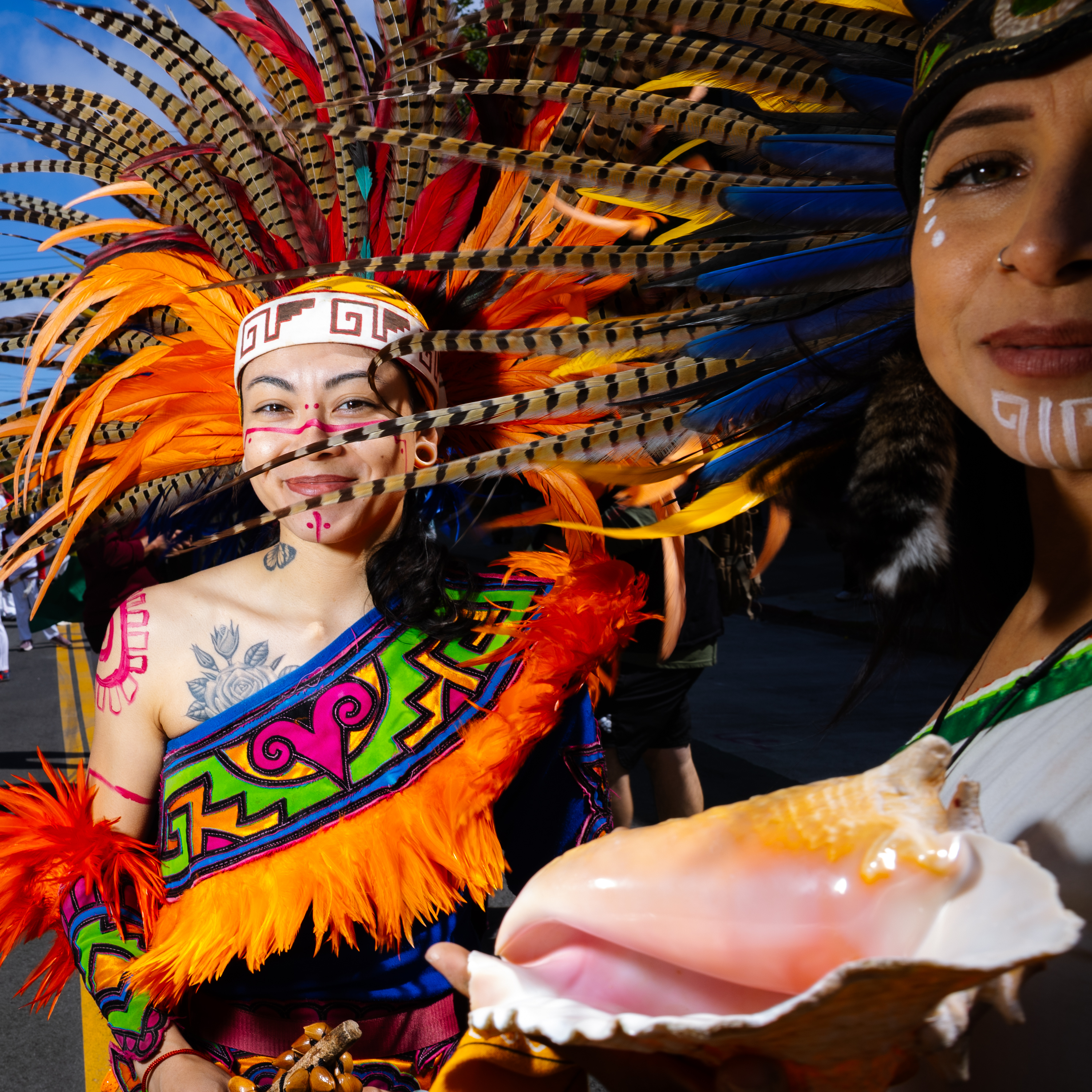 Two people in vibrant traditional attire, adorned with colorful feathers and face paint. One holds a conch shell. The background is a clear blue sky.