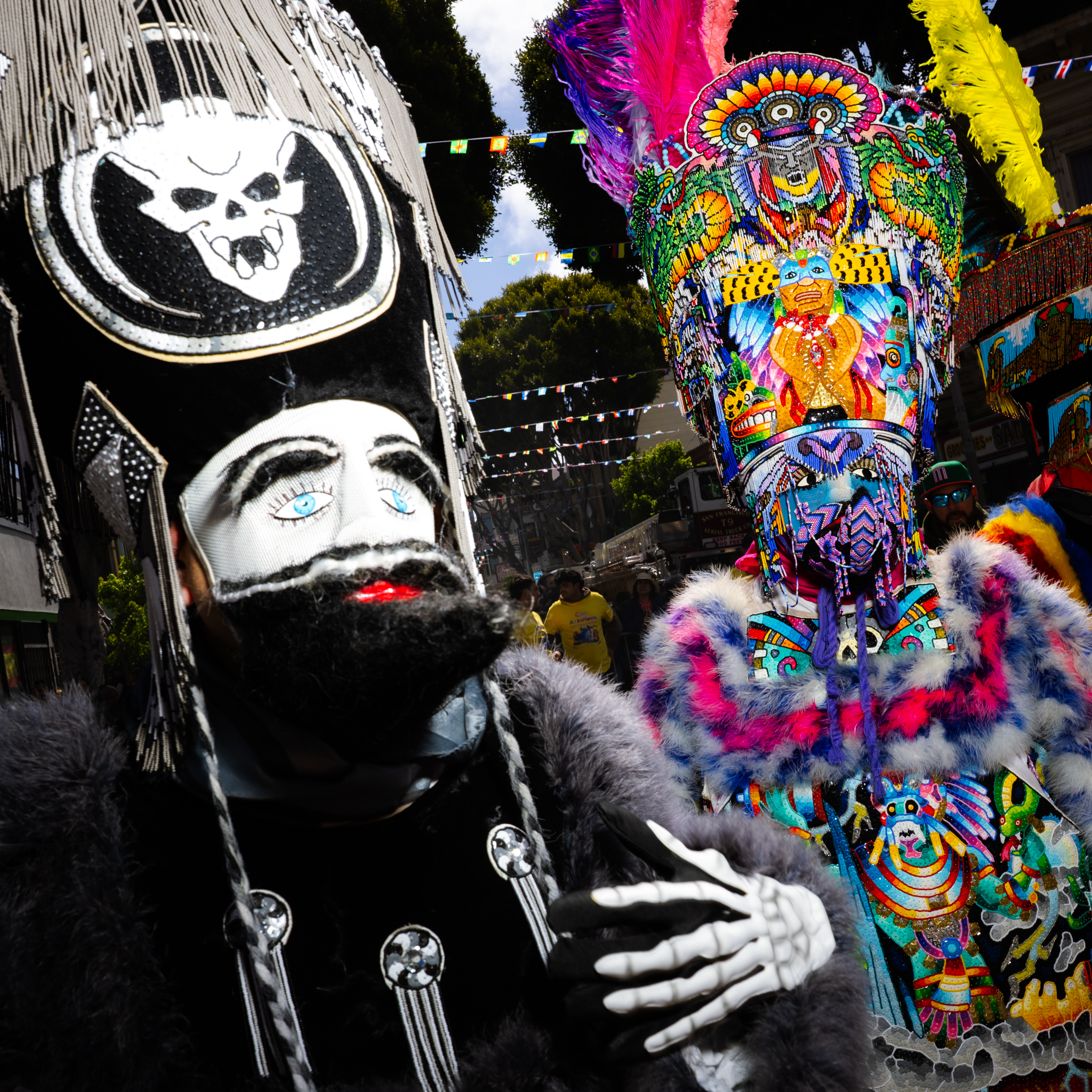 Two people wear elaborate, colorful costumes with ornate headpieces, one depicting a skull. They are in a festive street setting with flags above.