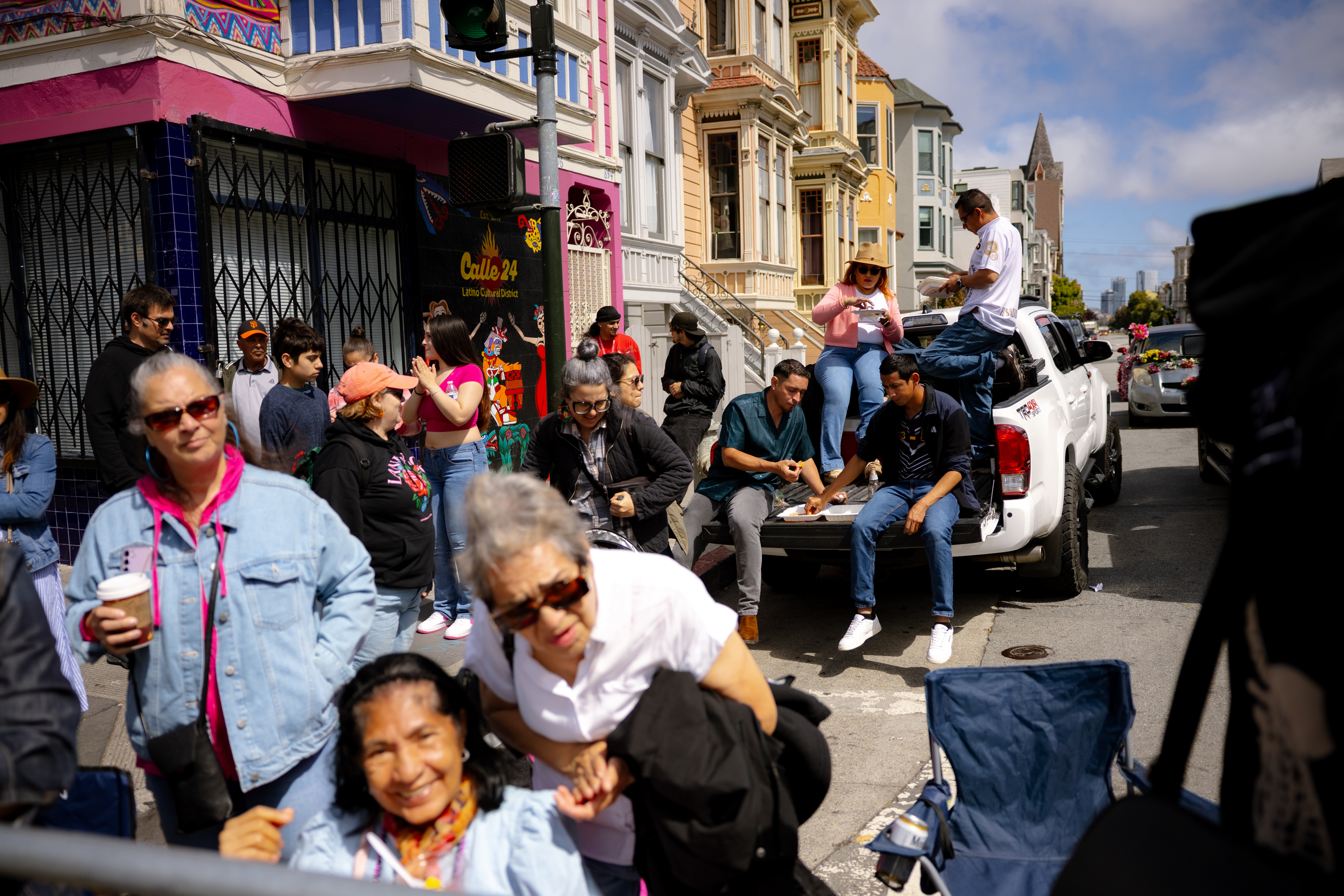 A lively street scene shows people gathered on a colorful urban sidewalk. Some are sitting on a white truck's tailgate, eating and chatting, while others are standing nearby.