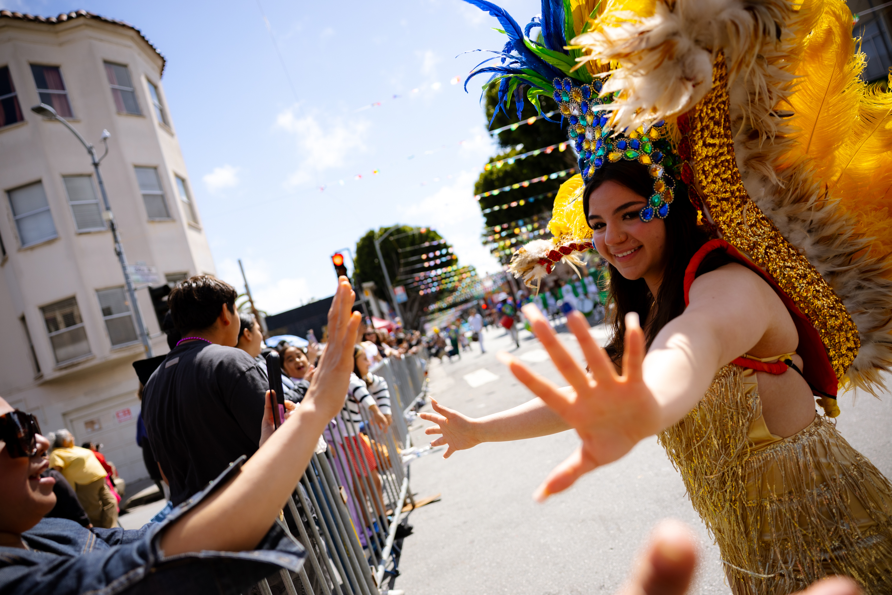 A smiling person in vibrant feathered attire high-fives people along a parade route, with festive decorations and a colorful urban backdrop.