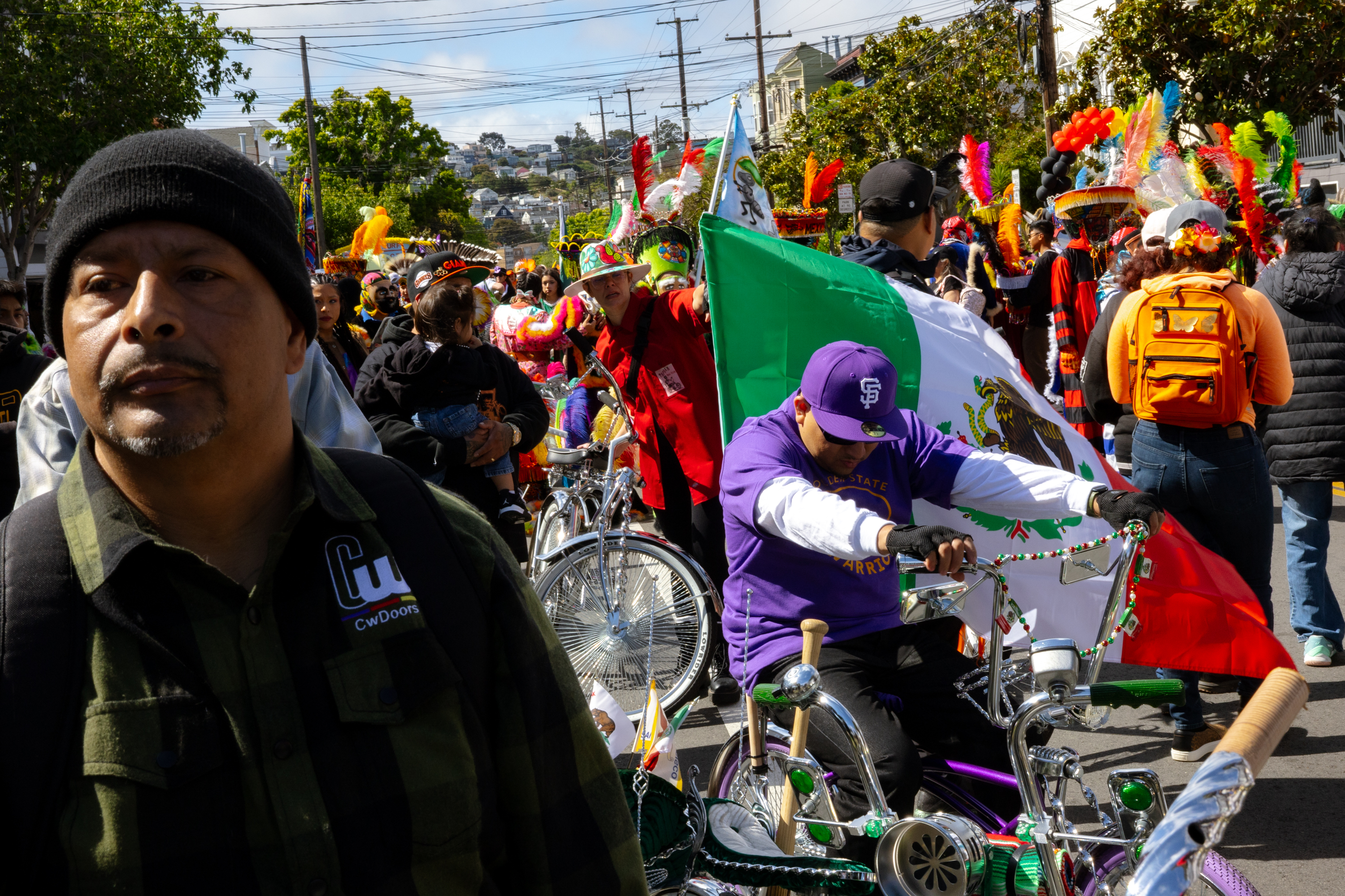 A colorful street parade features people in festive attire, with a man on a bike holding a Mexican flag. Vibrant decorations and a lively crowd fill the scene.