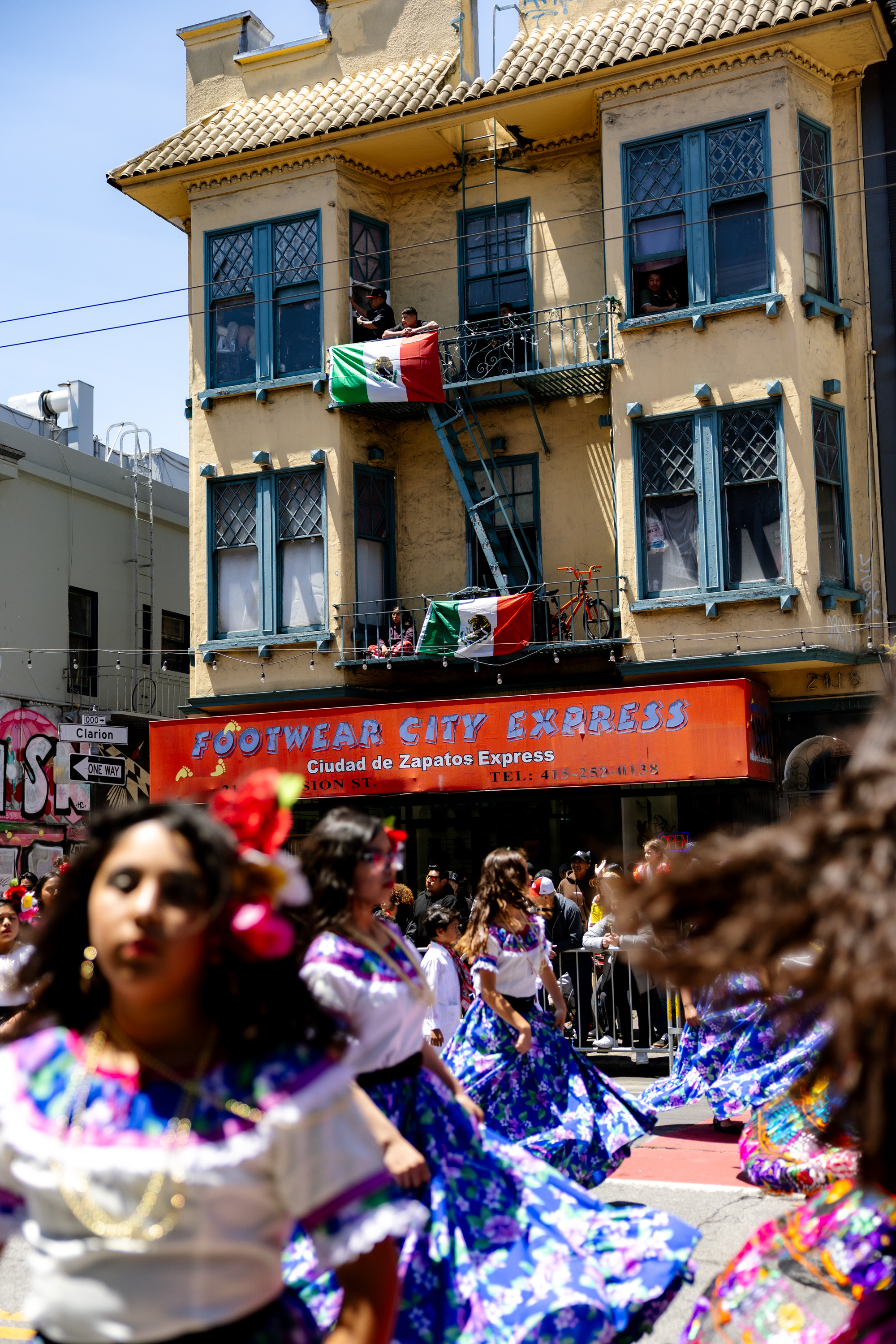 The image shows women in colorful traditional dresses dancing in a street parade. Behind them is a building with Mexican flags and a sign &quot;Footwear City Express.&quot;