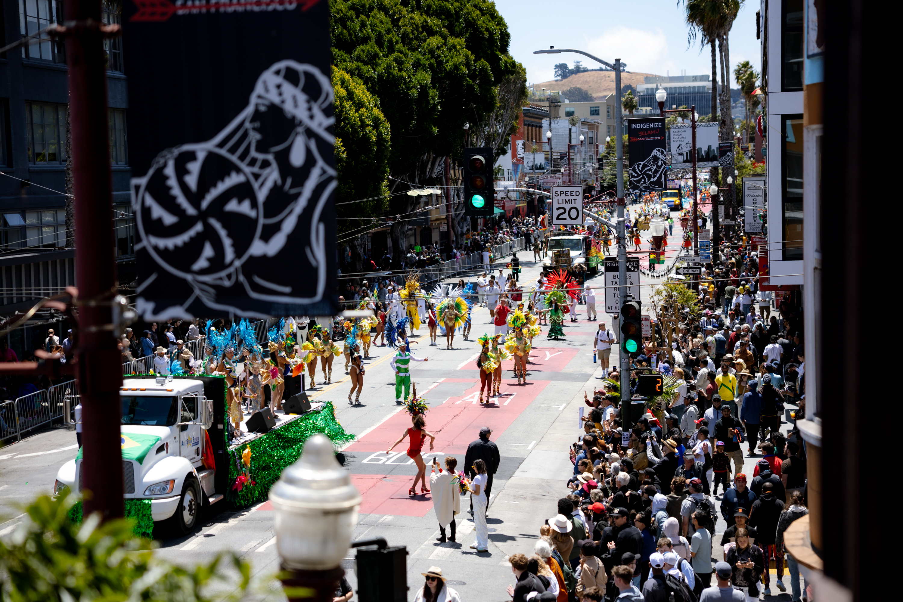 A vibrant parade fills the street with colorful costumes and a large crowd. Performers in bright outfits march alongside a decorated truck under sunny skies.