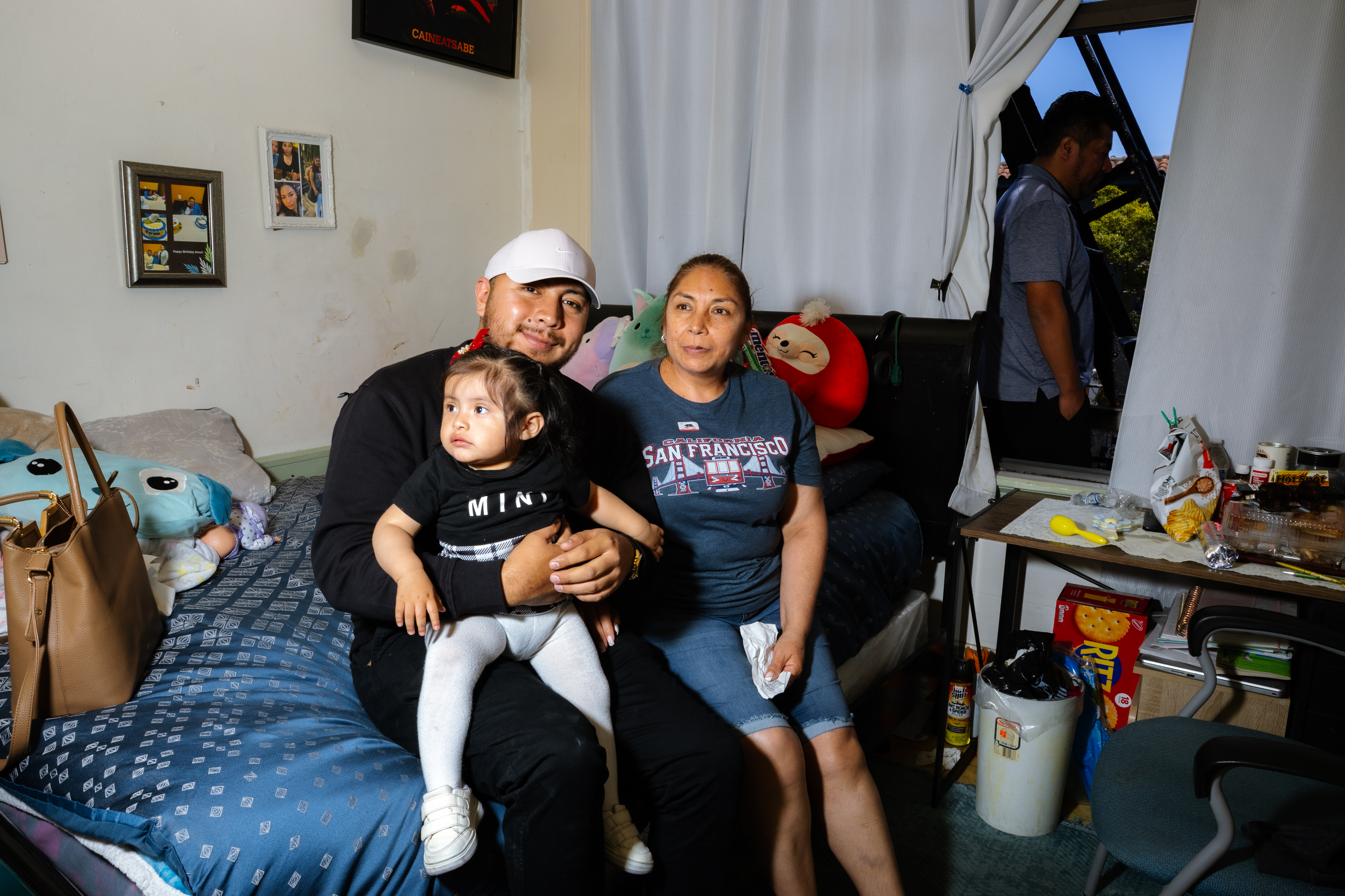 A family of three poses on a bed with a child sitting on an adult's lap. There's a stuffed toy behind them, a cluttered table, and a person near the window.